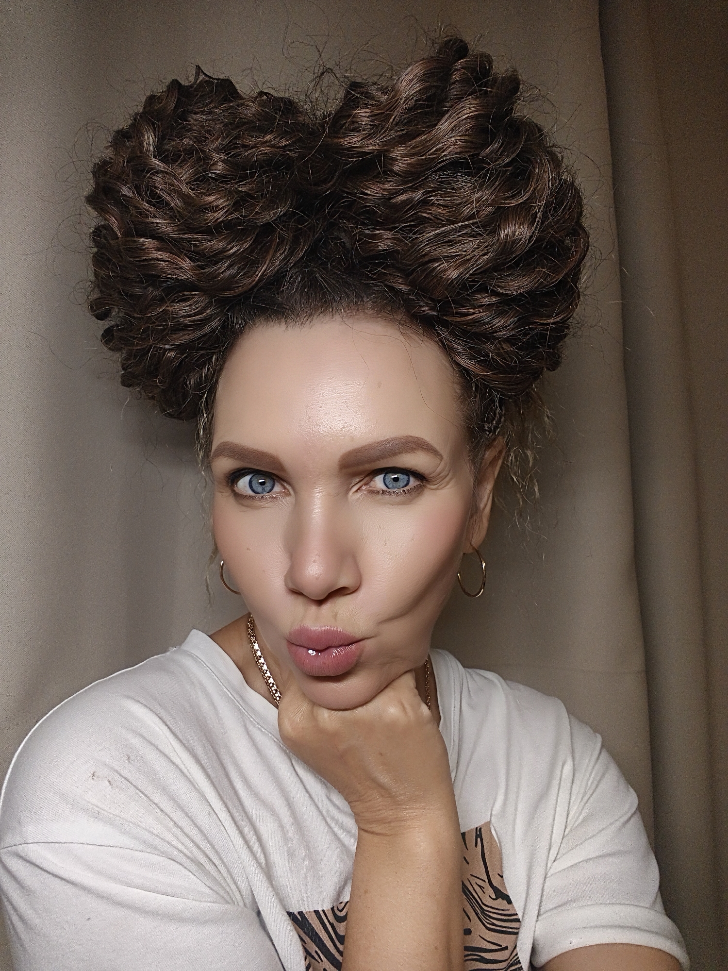 An adult female subject is depicted in a close-up, head-and-shoulders portrait shot within an indoor environment in Unknown, Unknown. She possesses voluminous, intricately curled dark brown hair styled high on her head, forming two distinct, rounded masses above her forehead. Her fair-skinned face features bright blue eyes looking directly into the camera, neatly defined light brown eyebrows, and full, glossy pink lips pursed into a pronounced pout. She wears small gold hoop earrings and a thin gold chain necklace. Her right hand is positioned under her chin, with her thumb visible. She is dressed in a white short-sleeved t-shirt, a portion of which displays a black and tan animal print pattern on the lower right side. The background consists of plain, light beige fabric or wall surfaces, softly illuminated.