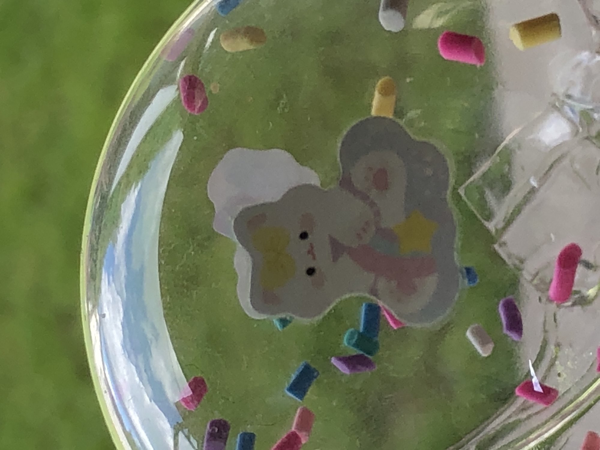 A clear spherical object filled with colorful confetti and a sticker of a white cat with a yellow bow is visible against a blurred green background. The cat sticker is positioned in the center of the sphere and is depicted with a pink ribbon or treat in its paws, on a cloud shape with a yellow star. The confetti includes rod-shaped pieces in various colors such as pink, blue, purple, and yellow, as well as some round pink pieces. The sphere's surface reflects the sky, showing blue with white clouds. The background appears to be an outdoor setting with green grass.