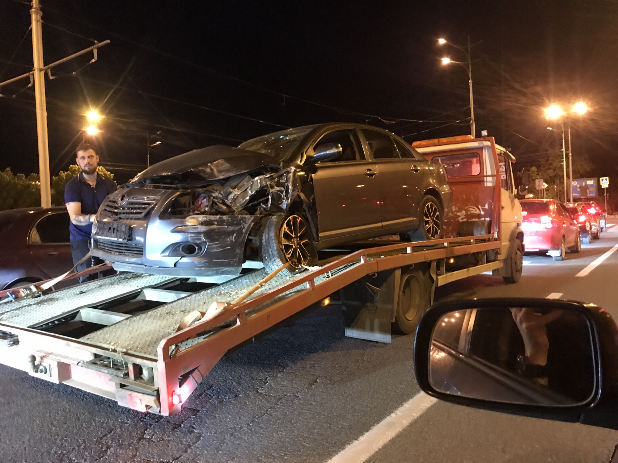 The image captures a nighttime scene on a road, where a severely damaged dark grey or silver sedan is being loaded onto or has already been loaded onto a flatbed tow truck. The front end of the sedan is heavily crumpled, with the hood bent upwards, the bumper broken, and the grille and headlights extensively damaged. The car features distinctive black and orange/gold spoked wheels.
A man with a beard and tattoos on his arms, wearing a dark blue polo shirt, stands on the left side of the frame, next to the front of the damaged car. He appears to be observing the process. The tow truck is red or orange, with an amber light bar visible above its cab. Behind the tow truck, a queue of red cars, possibly in traffic, stretches into the distance. Another dark vehicle is partially visible on the far left, next to the man.
The scene is illuminated by multiple streetlights, casting bright halos and reflections. Utility poles with overhead wires are visible against the dark sky. The road surface is dark asphalt with a clear white line marking the edge in the foreground. The photograph is taken from inside another vehicle, with its black side mirror reflecting a partial view of what appears to be an arm or hand in the bottom right corner. No clear readable text is visible on any signs or vehicles.