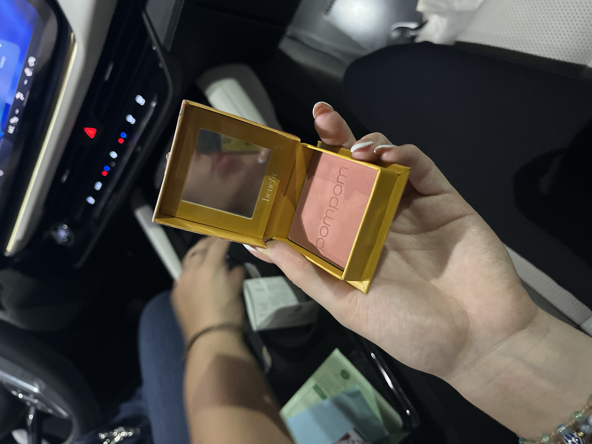 A person's right hand holds an open, yellow makeup compact inside a car. The compact features a small mirror on the left side, displaying the brand name "Benefit", and a rectangular pan of pink blush on the right, labeled "pompom" vertically. The hand has light skin and manicured nails with white tips. A multi-colored beaded bracelet is visible on the wrist of the person's other arm, which is partially seen in the background alongside a leg dressed in blue jeans.
The setting is the dark interior of a vehicle, likely at night or in low light, indicated by the illuminated dashboard controls. To the left, a digital car screen glows blue, and below it, a control panel shows various buttons, including a prominent red triangular hazard light button and several smaller white, blue, and red indicator lights. The overall scene is dimly lit, focusing attention on the compact and the hand holding it. The provided location context for this media is San Bernardino Tlaxcalancingo, Mexico.