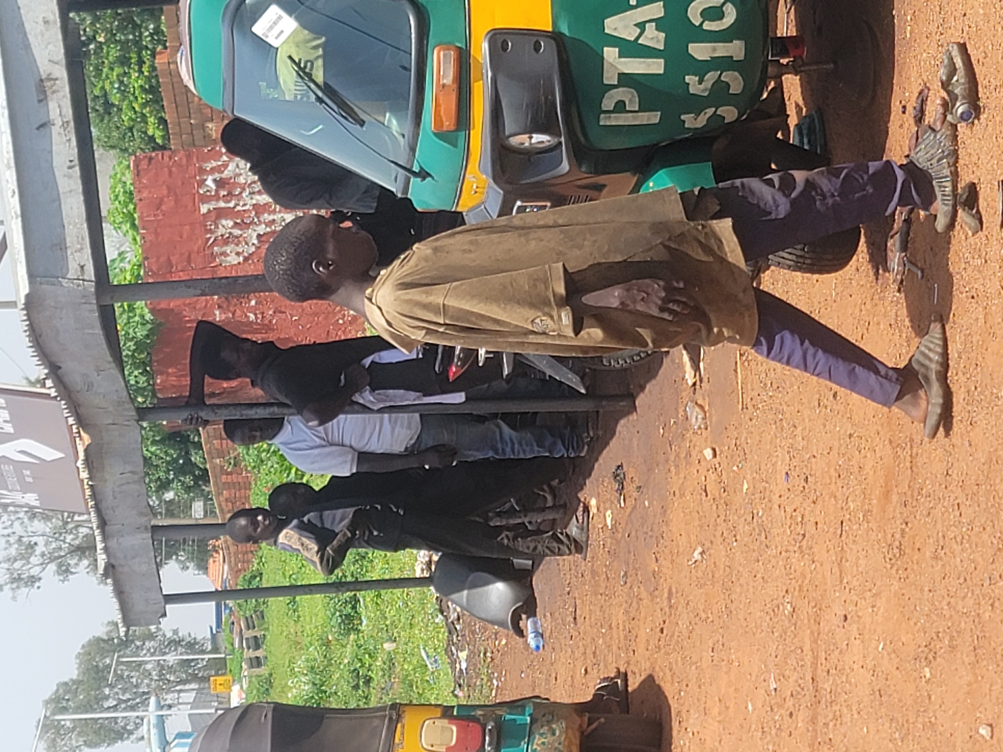 A group of people are gathered around a green and yellow motorized tricycle, commonly known as a "tuk-tuk." The scene appears to be outdoors, with a dirt ground and some greenery in the background, suggesting a rural or semi-urban setting in Barakin Lerawi, Nigeria. The lighting indicates it is daytime, likely sunny.
The main subject in the foreground is a young boy wearing a light brown, loose-fitting shirt and dark pants. He is standing with his hands on his hips, looking towards the viewer with a neutral expression. Behind him, other individuals are also present, some appearing to be waiting or observing. One person, wearing a white t-shirt, is visible near the tricycle. Another person, dressed in dark clothing, stands beside the boy.
The motorized tricycle has text on its side in white lettering against a green background. Partially visible words include "PTA" and "SSIC." This suggests the tricycle may be associated with a PTA (Parent-Teacher Association) or a similar organization. Other objects in the scene include a water bottle lying on the ground near the tricycle and what appears to be debris scattered on the dirt.
The overall impression is a snapshot of everyday life, possibly at a gathering point for transportation or a community event. The focus on the people and the common mode of transport provides a glimpse into the local environment.