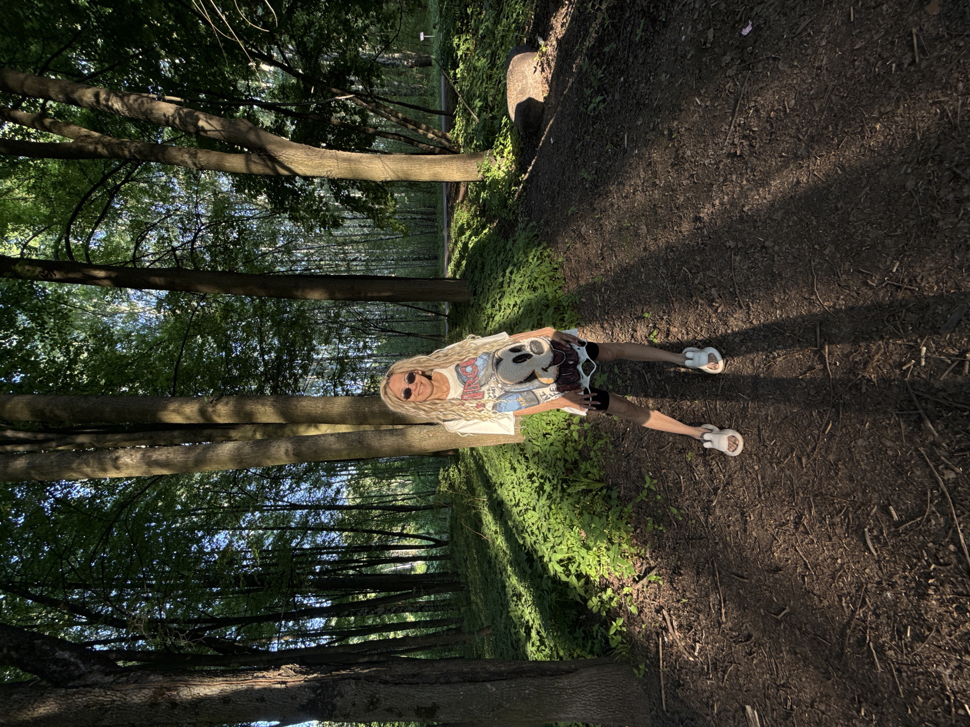 The image shows a blonde woman standing in a forest. The location is Шебунино, Russia. She is wearing a t-shirt with a cartoon graphic and sandals. The background consists of trees, foliage, and a dirt path. Sunlight is filtering through the trees.