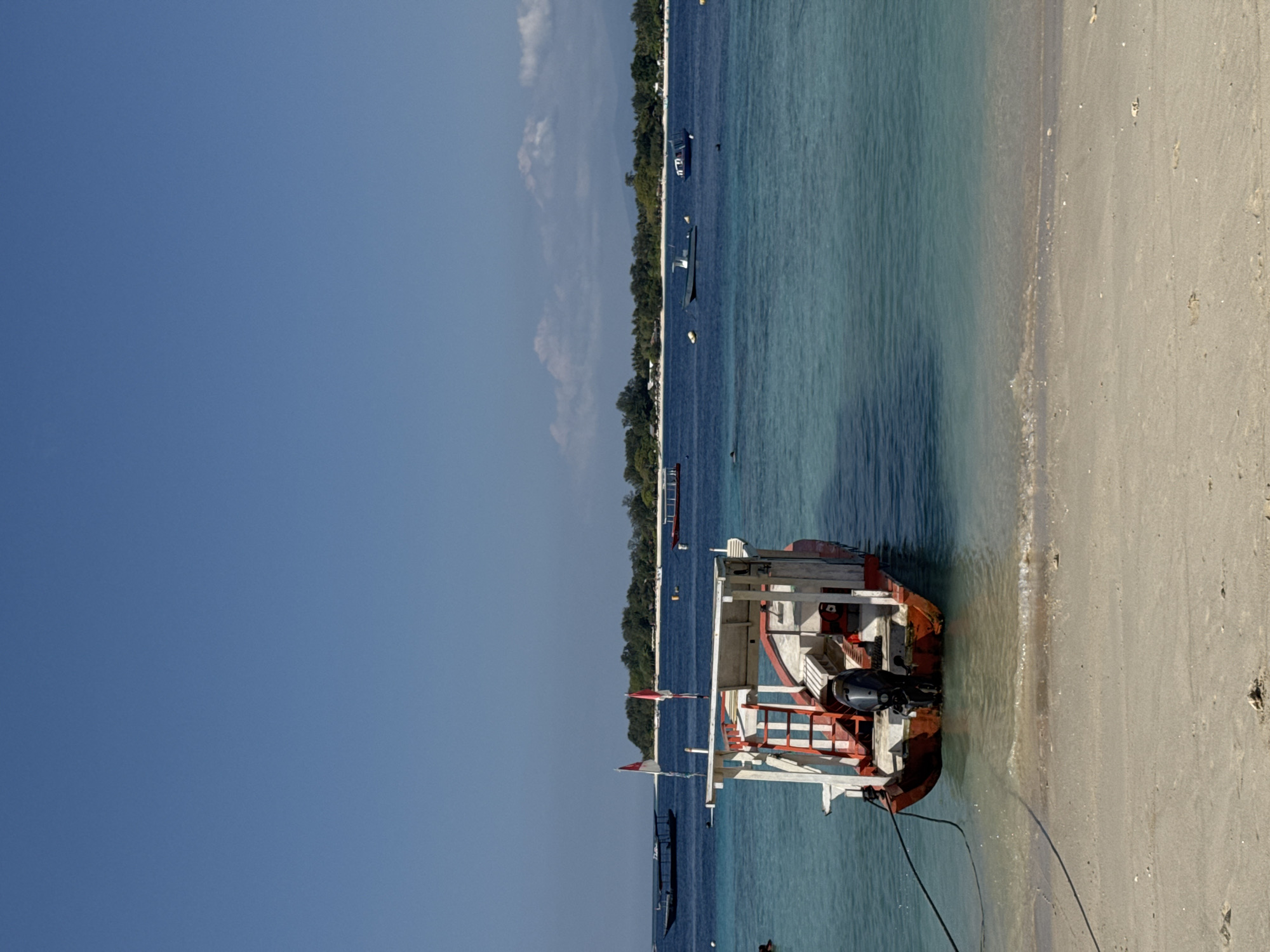 The image depicts a tropical beach scene in Indonesia. A small, red and white boat with an outboard motor is anchored close to a sandy shore. The water is clear and turquoise, with gentle waves lapping the beach. In the background, a lush green island with trees and buildings is visible across the sea, under a clear blue sky. Several other boats are scattered across the water, and a few people are swimming or wading near the shore. The overall atmosphere is serene and idyllic.