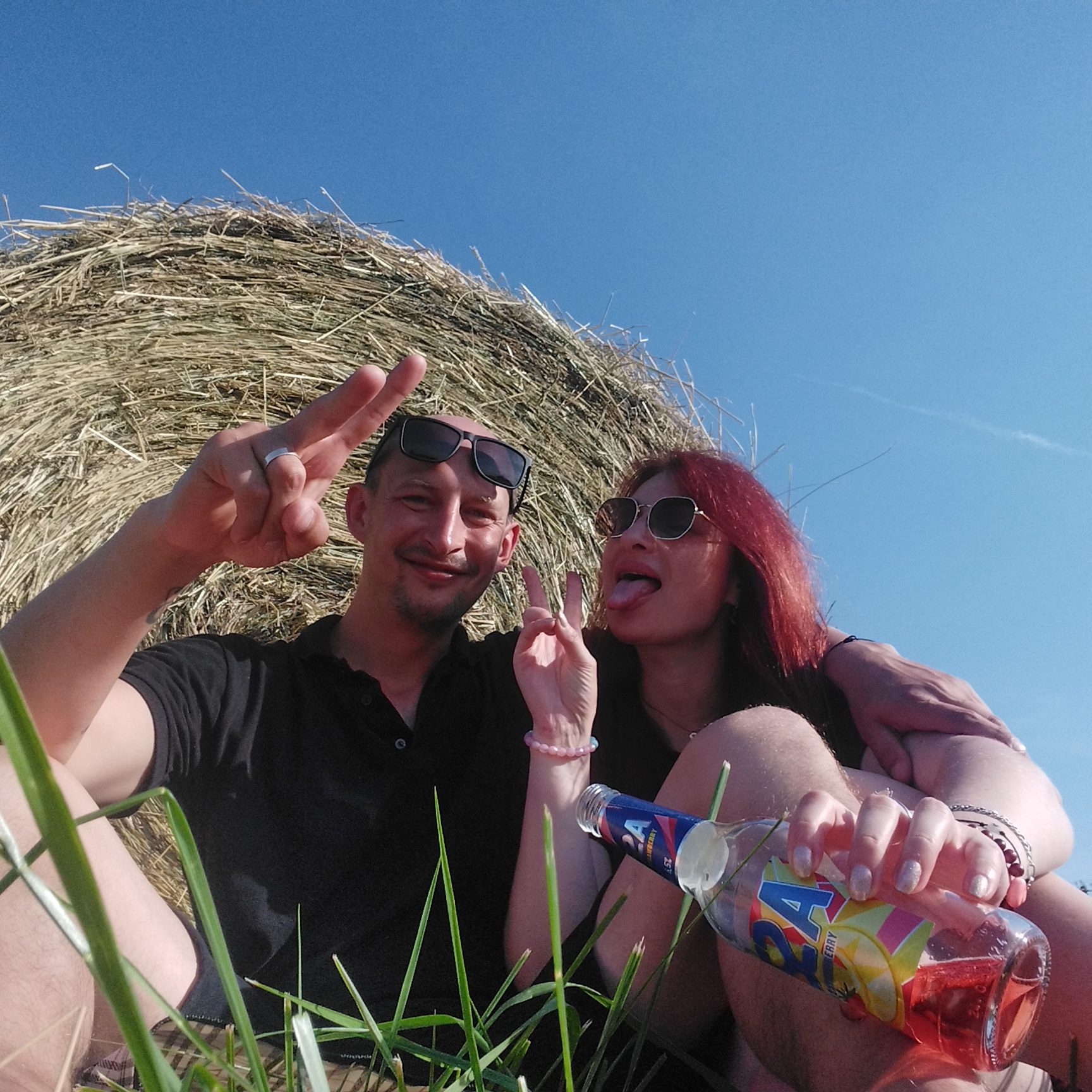 In an outdoor setting, a man and a woman are captured in a selfie, seated in front of a large haystack under a clear blue sky. The man, on the left, wears a black polo shirt and sunglasses perched on his head. He flashes a peace sign with his right hand, revealing a ring on his left ring finger. The woman, to the right, sports red hair and sunglasses. She playfully sticks out her tongue and also makes a peace sign with her left hand. A partially visible bottle of a pink beverage lies between them, held by the woman. Green grass and stalks are in the foreground, partially obscuring the couple. The haystack dominates the background, its dry, textured surface contrasting with the smooth blue sky. The scene appears to be set in a rural or open field location, far from any discernible city infrastructure.