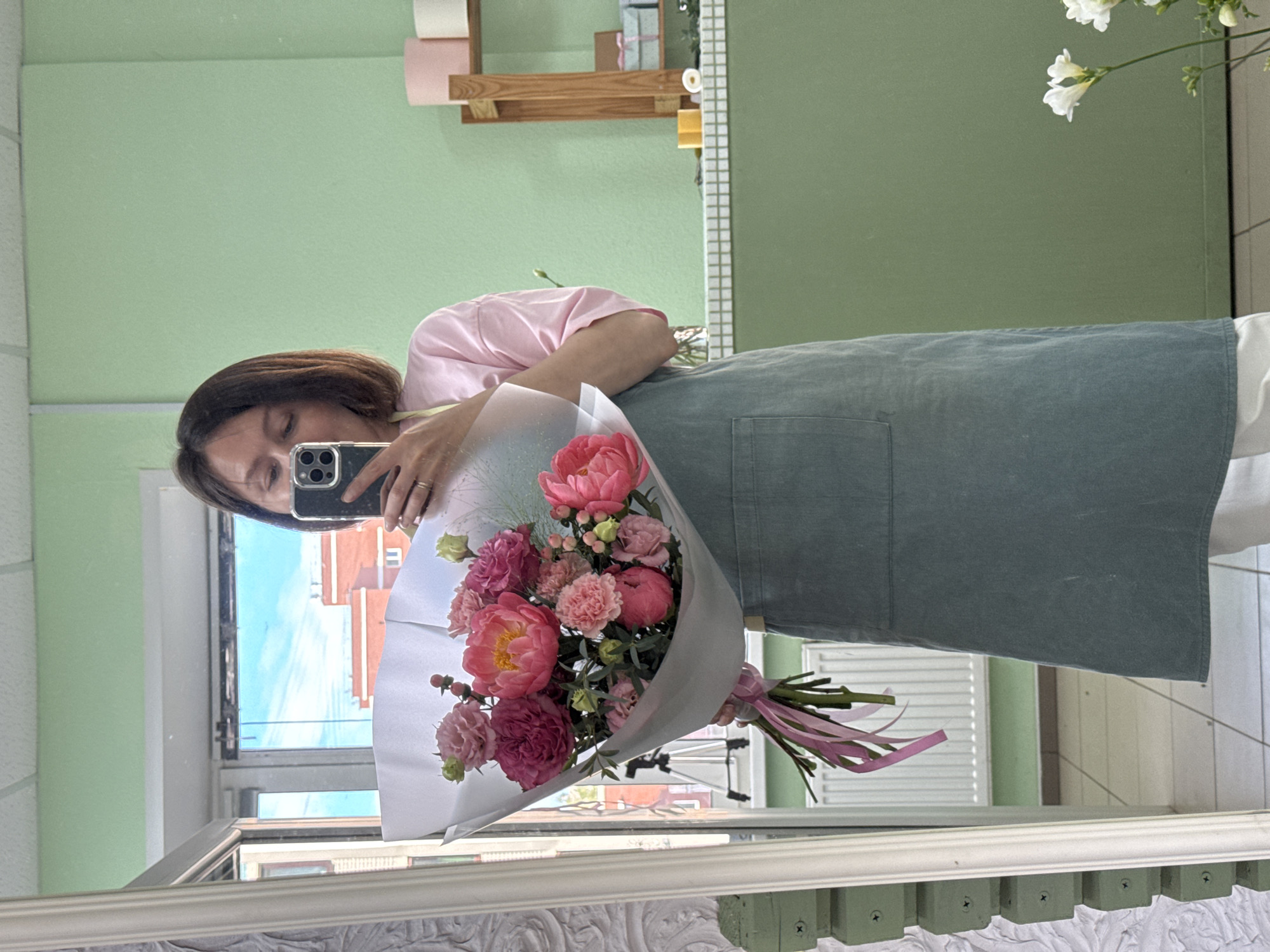 A woman is taking a selfie in a mirror, holding a bouquet of pink and coral flowers. She is wearing a pink t-shirt and a teal apron. The background appears to be a flower shop, with shelves and decor visible. The reflection in the mirror shows a window with a view of buildings and a blue sky. The image was taken in Izhevsk, Russia.