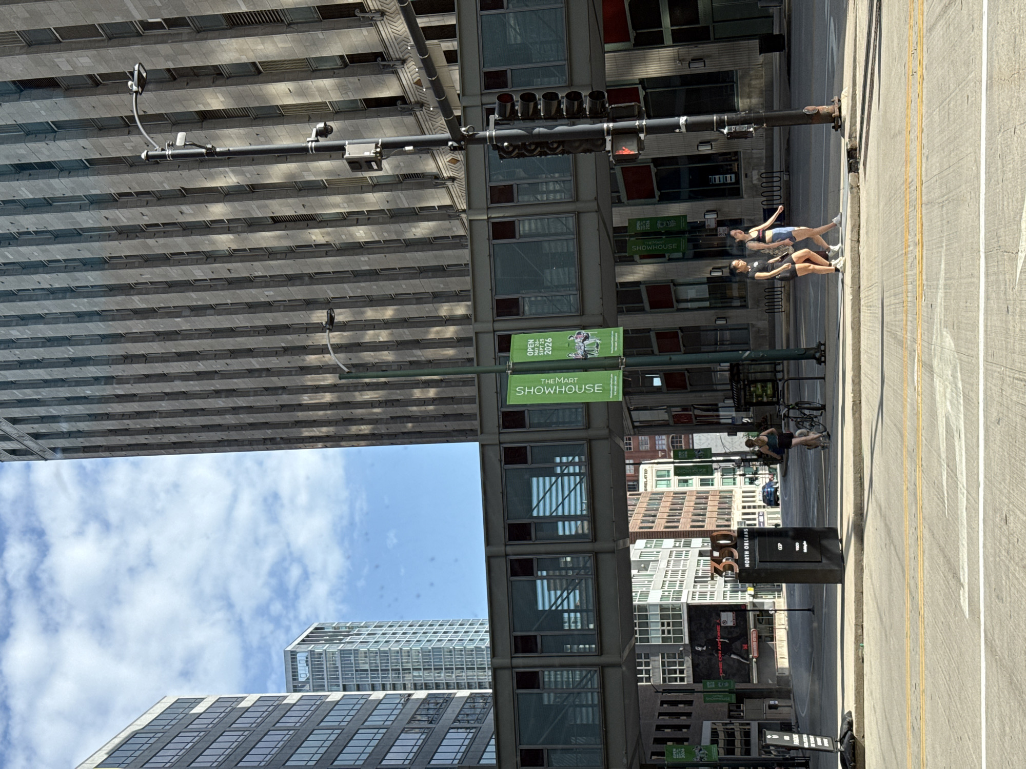 In Chicago, Illinois, United States of America, on a sunny day with scattered clouds, a street-level view captures urban architecture and pedestrian activity. The scene is dominated by tall buildings, including a modern skyscraper with reflective glass windows and a more traditional stone-clad building. A skybridge connects two of the structures, featuring large glass panels. In the foreground, a street with lane markings is visible.
Several people are present. Two young women are walking on the sidewalk, dressed in athletic attire. Another woman is walking with a bicycle next to her. On the sidewalk, a black kiosk displays "350 NORTH ORLEANS" and other text. Green banners advertising "THEMART SHOWHOUSE" hang from the skybridge. A traffic light, pole-mounted camera, and streetlights are also visible. The overall environment suggests a bustling downtown area.