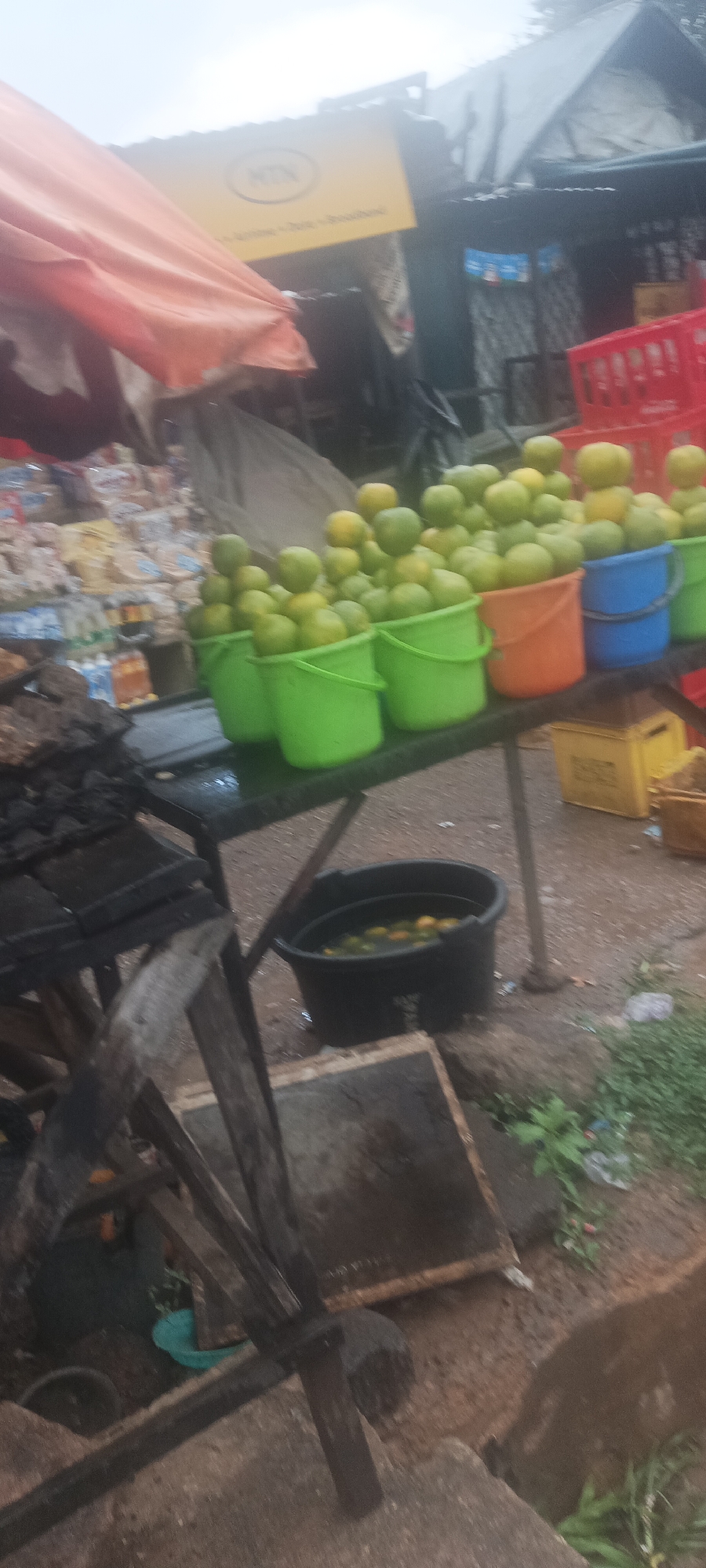 The image depicts a roadside market stall in Kuru Dungus, Nigeria. The stall features a table laden with green and yellow citrus fruits, likely oranges or tangerines, displayed in colorful plastic buckets (green, orange, blue). A black bucket containing fruits soaking in water sits below the table. To the left, there's a charcoal grill and a table with various packaged food items. An MTN (mobile telecommunications network) sign is visible in the background, indicating a nearby business or service point. Red plastic crates, possibly for Coca-Cola, are stacked near the fruit. The environment appears humid, with a slightly overcast sky, and the ground is paved. No people are directly visible in the frame. The primary action is the display of produce for sale.