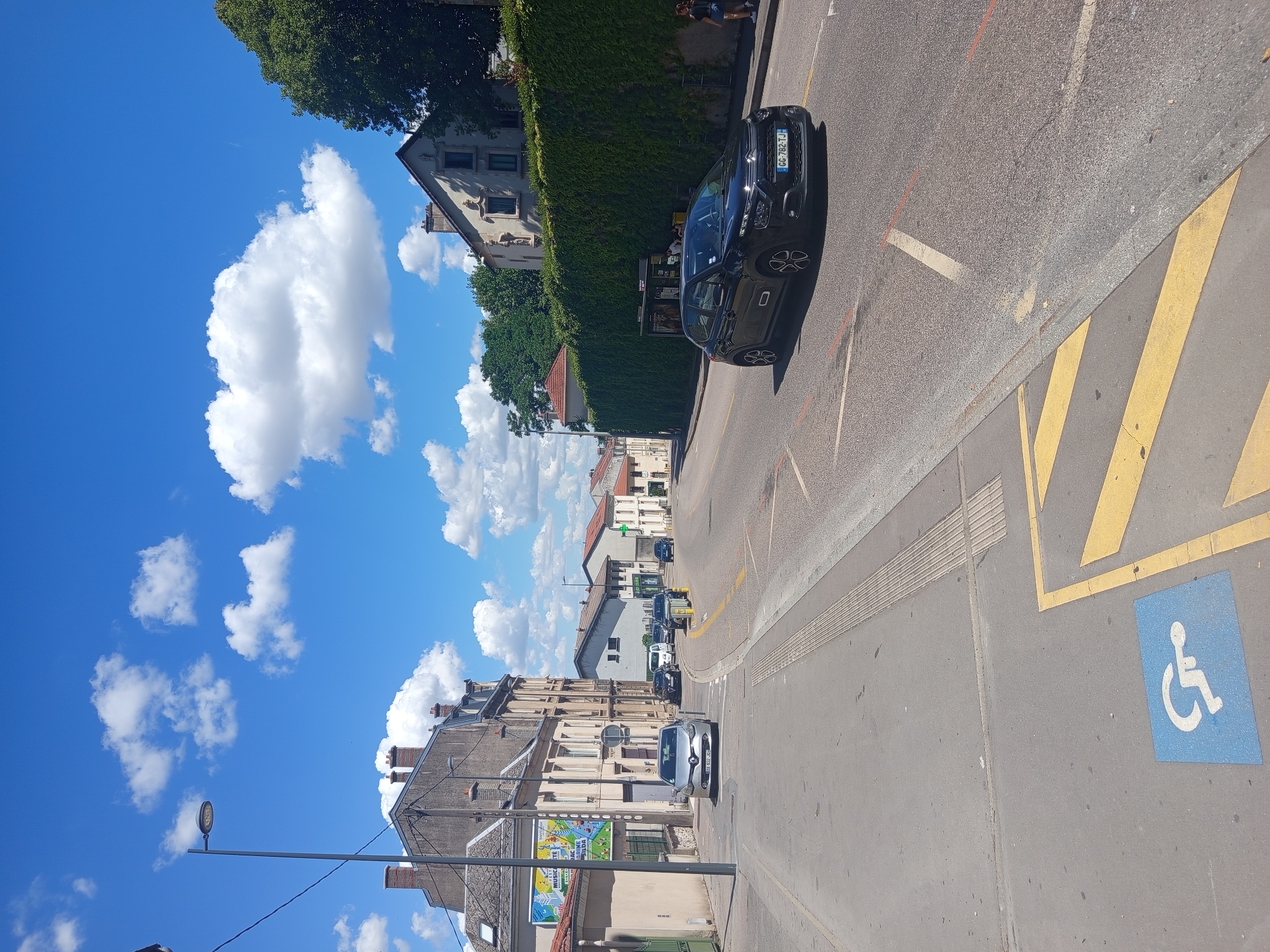 The image depicts a street scene in Nancy, France, under a bright blue sky with scattered white clouds. In the foreground, a blue square with a white wheelchair symbol indicates a disabled parking spot. Yellow diagonal lines are painted on the asphalt, forming part of a road marking. A dark-colored hatchback car, bearing the license plate "CG-782-TJ", is driving on the right side of the road. The street is lined with buildings on the left, including a multi-story building with a poster on its facade. On the right side of the road, a dense green hedge forms a wall. Further down the street, several other cars are parked or driving. A street light is visible on a pole on the left side of the image. The overall environment suggests a sunny day in a residential or commercial area.
