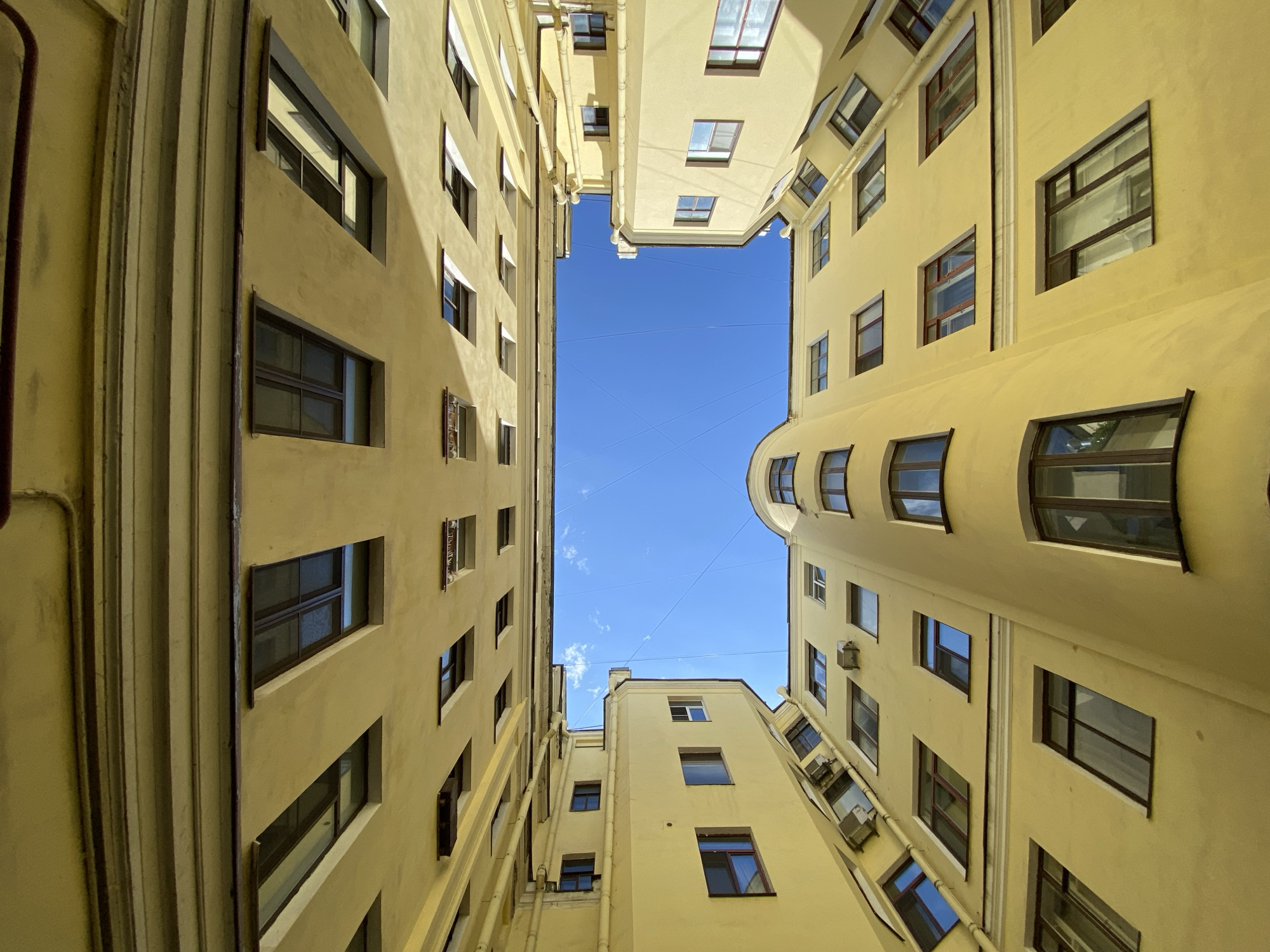 The image shows a view looking straight up from inside a courtyard surrounded by tall buildings. The buildings are uniformly painted in a light yellow or tan color. Numerous windows of various sizes punctuate the facades of the buildings. A small rectangle of bright blue sky is visible at the top of the frame, between the buildings. Cables stretch across the open sky.