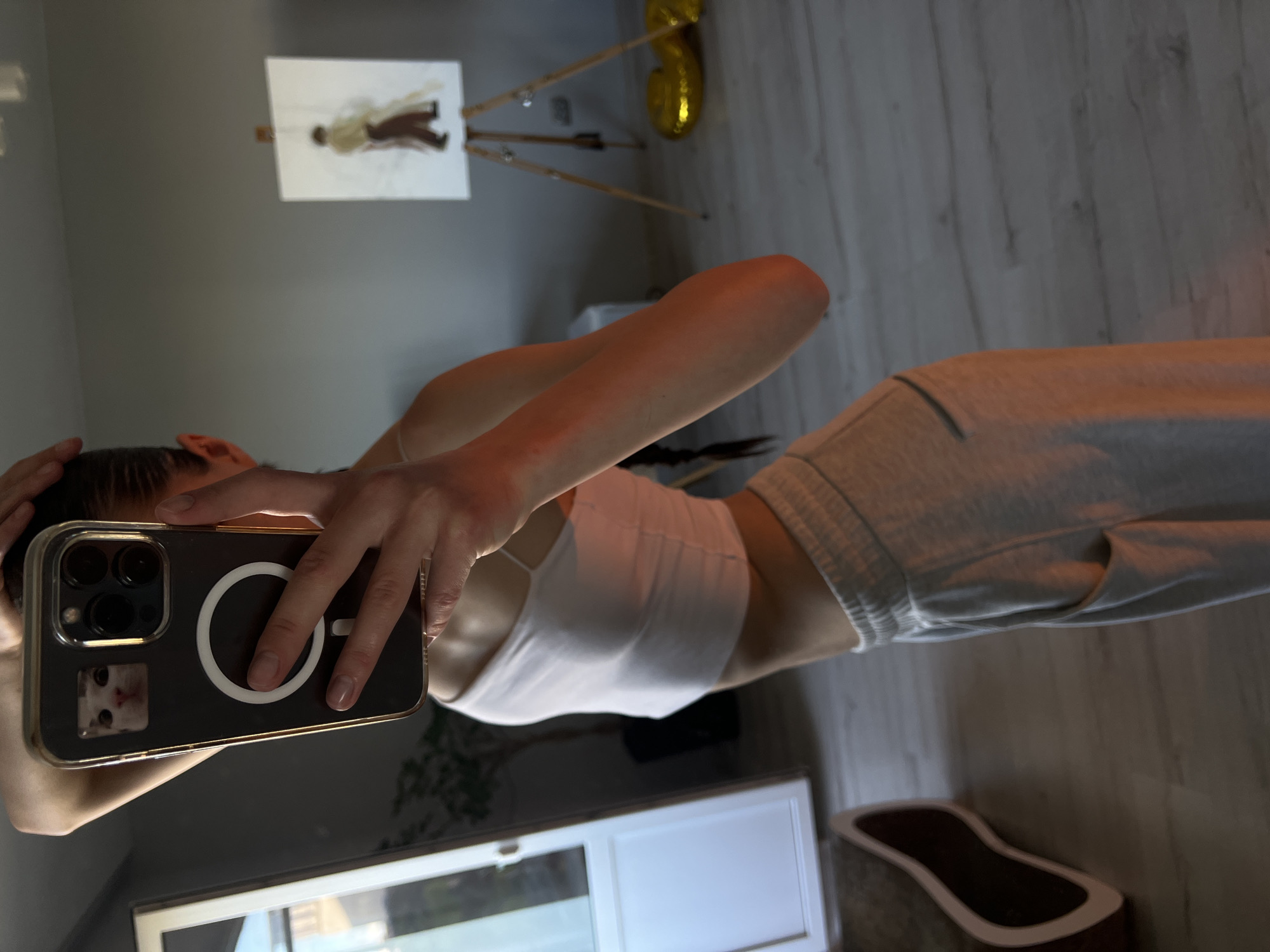 The image shows a young woman taking a selfie in a room. She is wearing a white crop top and grey sweatpants. Her hair is in a braid. In the background, there is a painting on an easel and some gold balloons. The room appears to be a living room with wooden floors and grey walls. There is no indication of the location being Ternopil, Ukraine.