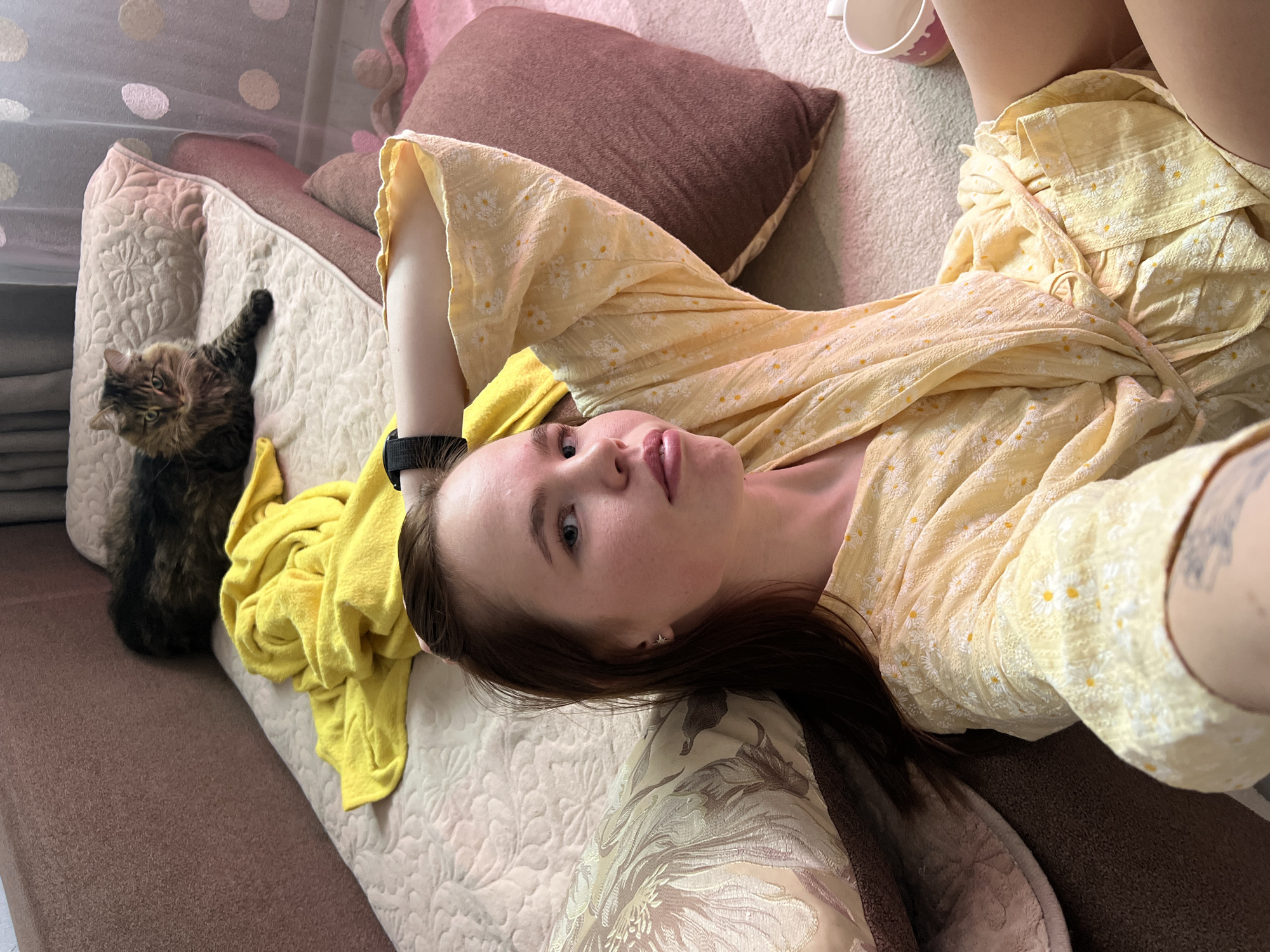 A young woman is lying on a sofa in what appears to be an indoor residential setting in Chaikovsky, Russia. She is fair-skinned, with dark hair, and wearing a yellow floral-patterned garment. Her left arm is raised, with a black wristband visible. A long-haired cat is sitting upright to her left on the sofa. A yellow sweater or blanket is crumpled beside the cat. A brown pillow is also present on the sofa. A cup with a pink design is positioned in the background on the sofa. The couch has a patterned beige fabric. The overall environment appears domestic and comfortable.