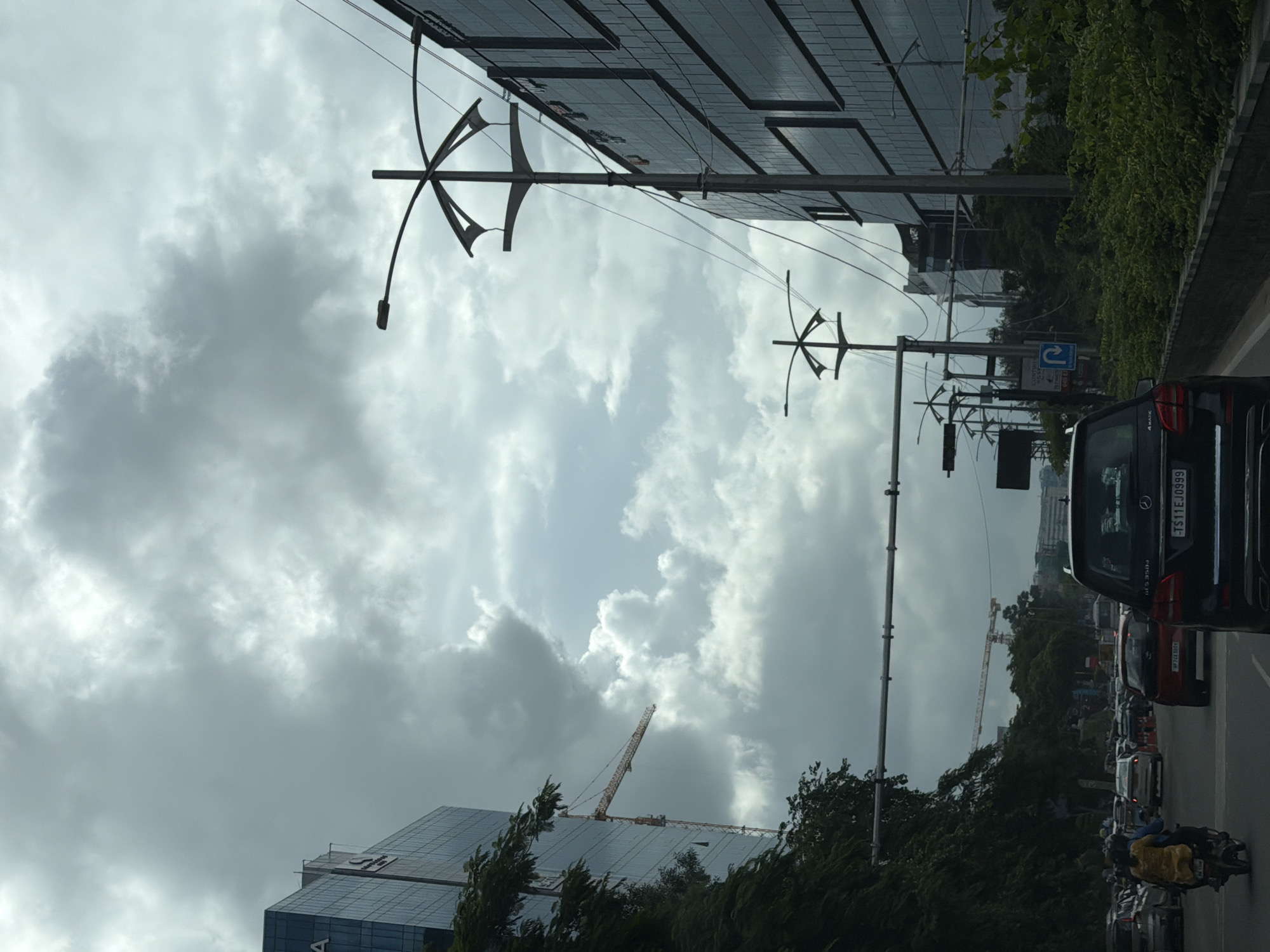 A low-angle, vertical shot captures a busy city street in Hyderabad, India, under a dramatic, overcast sky. Two modern, glass-fronted buildings flank the road, one on the left and a larger one on the right. A construction crane is visible in the distance, extending from behind the building on the left, suggesting ongoing development. The road is filled with traffic, including several cars, a motorcycle with two occupants, and the back of a black SUV prominently in the foreground with a visible license plate reading "TS11EJ0999." Traffic lights and street lamps are strategically placed along the roadway, with power lines crisscrossing the scene. Lush green foliage lines the side of the road, adding a touch of nature to the urban landscape. The atmosphere is heavy with the impending possibility of rain, indicated by the dark, thick clouds.