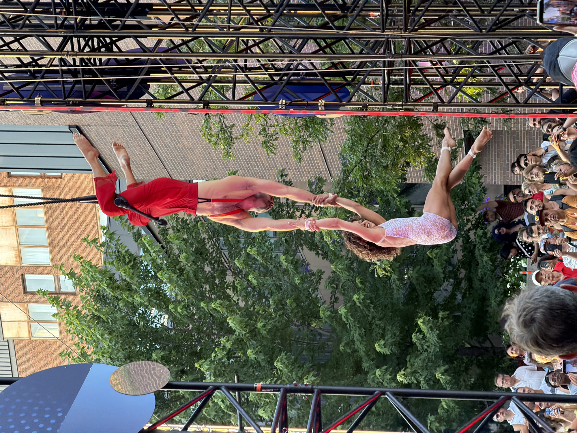 This is a vertical, eye-level, medium shot of an outdoor aerial performance at the Quartier des Spectacles in Montreal, Canada. Two performers, one male and one female, are suspended in the air, held by a taut rope. The male performer, on top, is wearing red trousers and suspenders and is upside down, gripping the female performer's wrists. He has fair skin and is looking downwards. The female performer, below, is wearing a white, sequined leotard and is in a bent-leg pose, facing left and looking up towards the male. Her hair is dark and curly. They are positioned against a backdrop of a brick building with many windows and lush green trees. To the right, a large metal truss structure, characteristic of outdoor event stages, is visible. A crowd of spectators is gathered at the bottom of the frame, many holding up phones to capture the performance. The lighting suggests it is daytime, likely late afternoon or early evening, given the warm light.