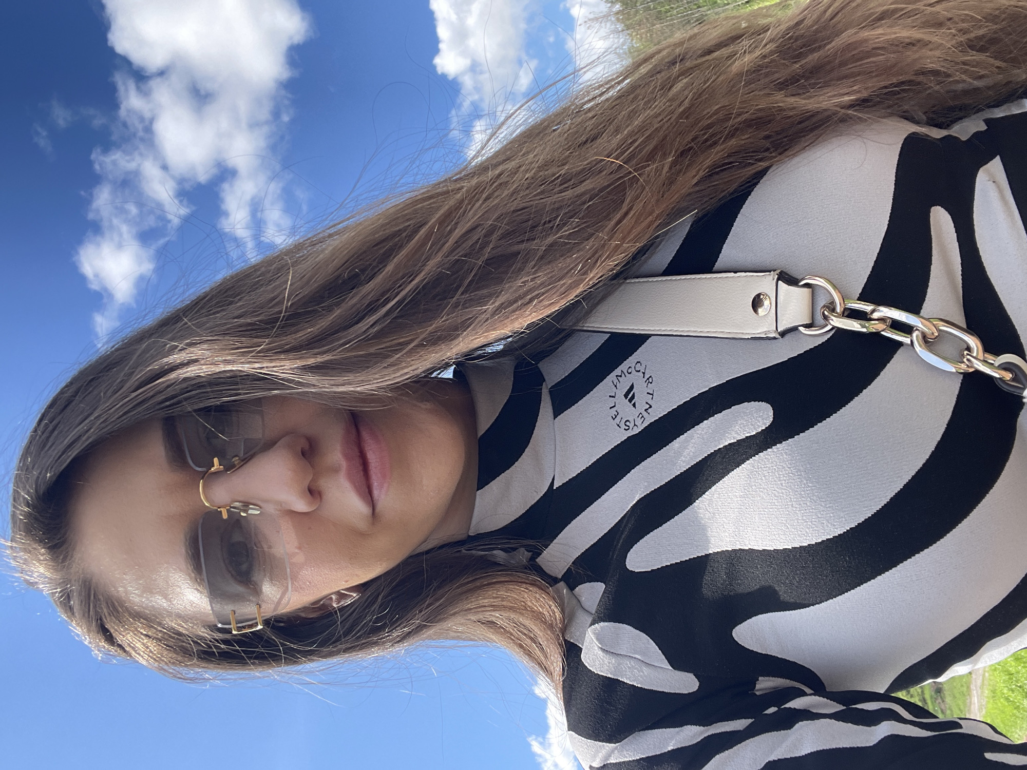 A young woman poses for a selfie outdoors. She is wearing rimless sunglasses and a black and white zebra-print turtleneck with the "Stella McCartney" logo. She has long brown hair and a white purse with a chain strap. The background is a bright blue sky with fluffy white clouds. Green grass is visible at the bottom of the frame. The picture was taken in Novo Selo, Bulgaria.