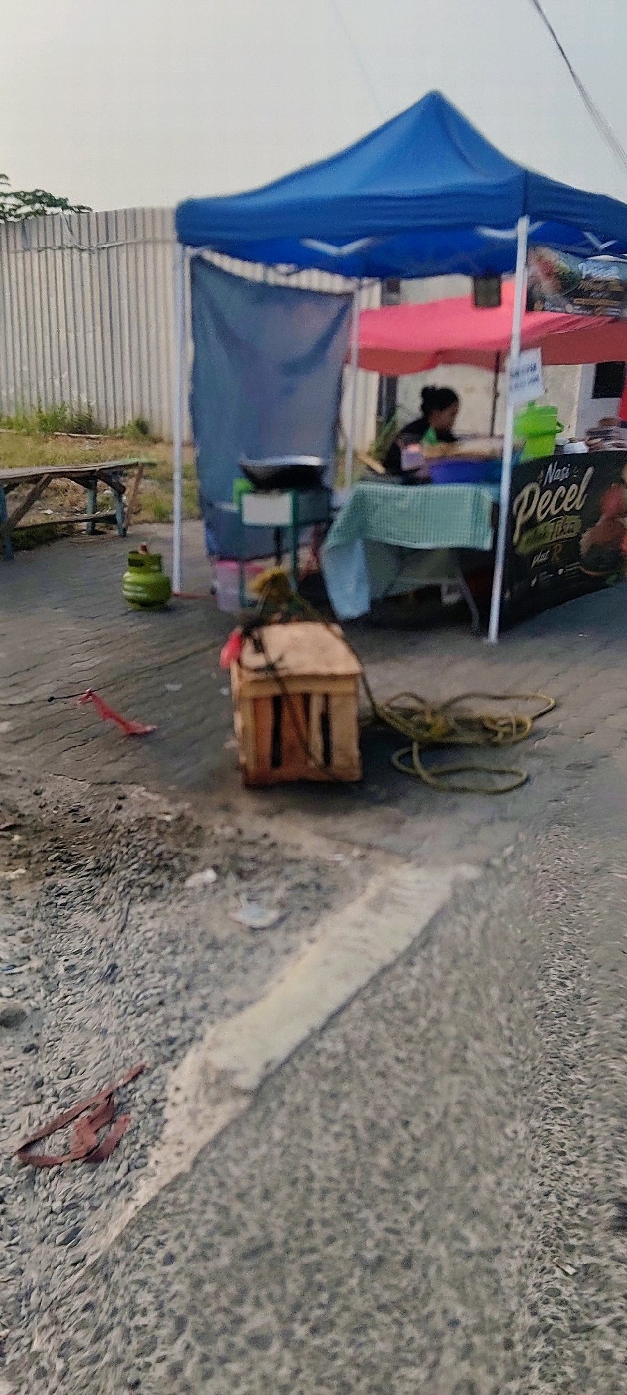 The image depicts an outdoor food stall operating under a blue and pink canopy, likely situated on a roadside in Mekarmukti, Indonesia. A woman, partially visible behind a table covered with a checkered cloth, is attending to the stall. Various food-related items are present, including what appears to be a wok on a stand and a sign that reads "Nasi Pecel." A green gas cylinder stands to the left of the stall, and a wooden crate with rope coiled on top is in the foreground. The ground is paved, and a metal fence is visible in the background, suggesting an urban or semi-urban environment. The overall lighting suggests it might be late afternoon or early evening.