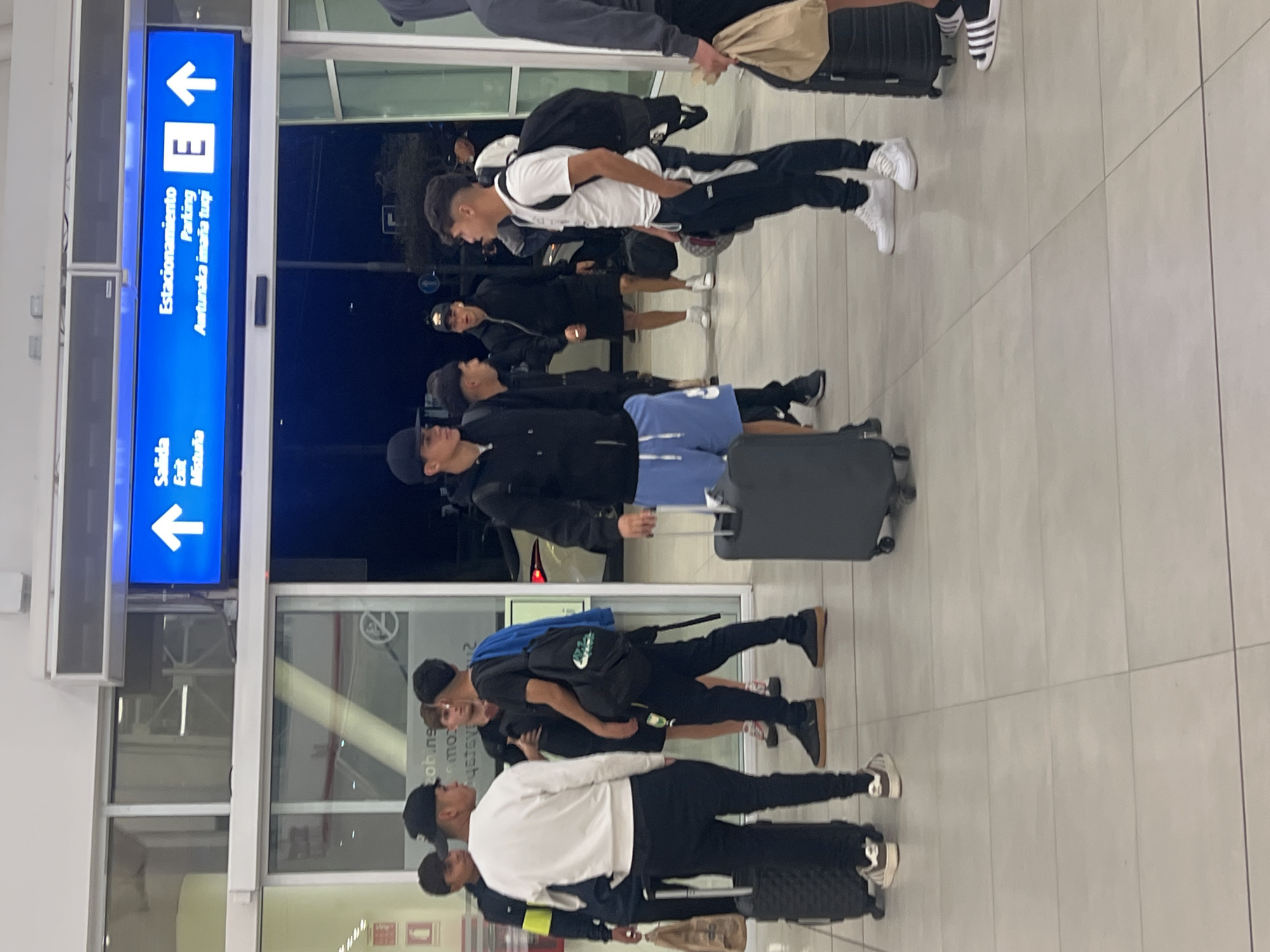 Here's a description of the image:
The image shows a group of young men, likely a team, standing in what appears to be an airport or transportation hub in Arica, Chile. They are positioned in front of a glass wall/entranceway. Many have luggage.
A blue sign indicating "Estacionamiento/Parking/Awtunaka imaña tugi" (Parking in Spanish and presumably a local language) and "Salida/Exit/Mistuña" is visible. The sign has directional arrows. The style of the sign and architecture suggest a modern building. The group appears to be waiting or possibly entering or exiting the building.
