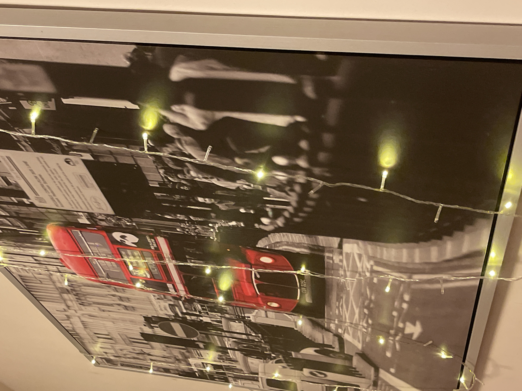 The image is a photograph of a framed artwork depicting a red double-decker bus in a black and white cityscape. Fairy lights are draped across the artwork, obscuring parts of the image with glowing orbs of light. The bus appears to be a classic Routemaster model, common in London. The background shows a street scene with buildings and a crowd of people. There is no indication of the city of Amman, Jordan in the image. The artwork is framed and mounted on a wall, with a portion of the wall and the edge of the frame visible.