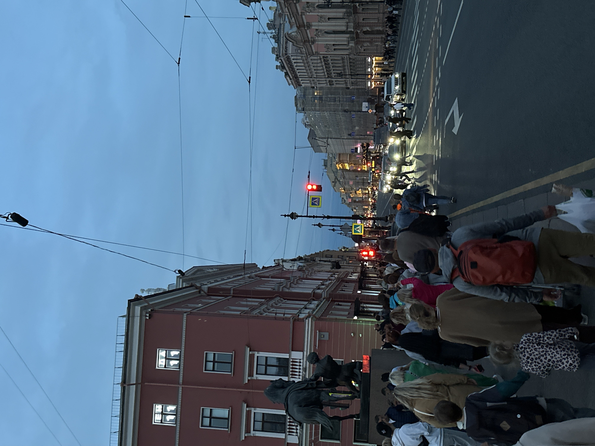 This is a vertical, eye-level, wide shot of a street scene in Saint Petersburg, Russia, likely in the evening given the dim, overcast sky and the artificial lights of the buildings and vehicles. A crowd of people are gathered on the sidewalk and crossing the street. They appear to be waiting or observing something, perhaps a parade or performance, as indicated by their unified direction of gaze towards the center of the street.
The architecture is ornate and historic, with multi-story buildings featuring classical detailing, suggesting a European city. The street is wet, reflecting the lights, implying recent rain or damp conditions. Cars are visible on the road, and a traffic light is red, indicating a halt in traffic. Pedestrian crossing signs are also visible. A statue of a horse and rider is partially visible in the foreground, adding to the historical context.
The people are dressed in casual, modern clothing. Their faces are not clearly discernible, but the overall atmosphere seems to be one of gathering and anticipation. There are no obvious signs of distress or overt celebration, but rather a sense of collective presence. The image captures a moment of urban life in a grand European city.