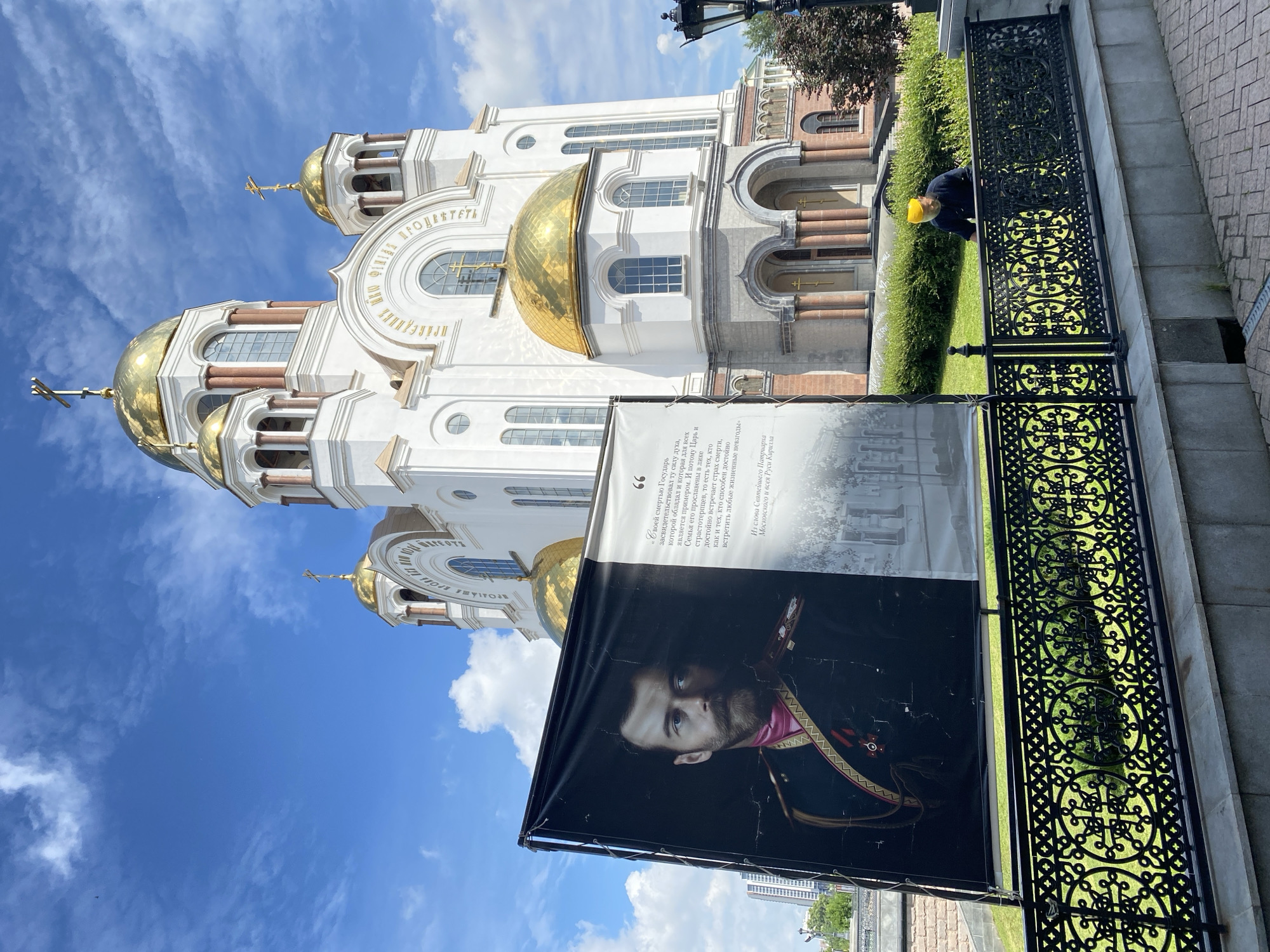 In Yekaterinburg, Russia, a banner featuring a portrait of Tsar Nicholas II is displayed in front of the Church on Blood, a prominent landmark built on the site where the imperial family was executed. The banner shows the Tsar in military uniform on the left, and text on the right, partially obscured. A person wearing a yellow hat is visible behind a decorative black metal fence in the foreground, tending to the grass. The sky is blue with scattered clouds, and the church, with its golden domes, is brightly lit by the sun. Lush green foliage surrounds the church and the area in front of it.