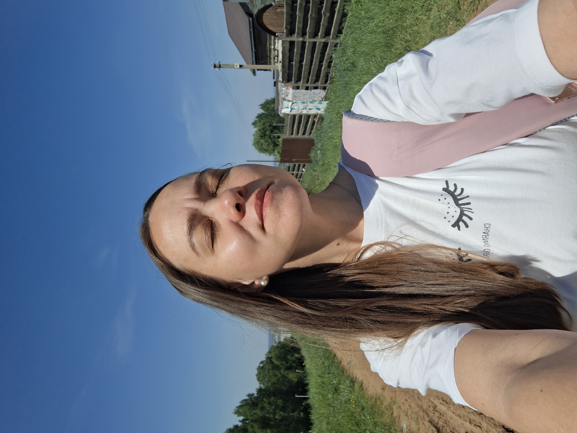 A woman is positioned in a photograph. She is wearing a white t-shirt with a graphic design, a pink long-sleeved shirt or covering, and an earring. She has closed eyes and brown hair. The background includes a bright blue sky, green vegetation, a wooden structure, and possibly a power pole. The location appears to be rural, possibly outdoors in Chaikovsky, Russia.