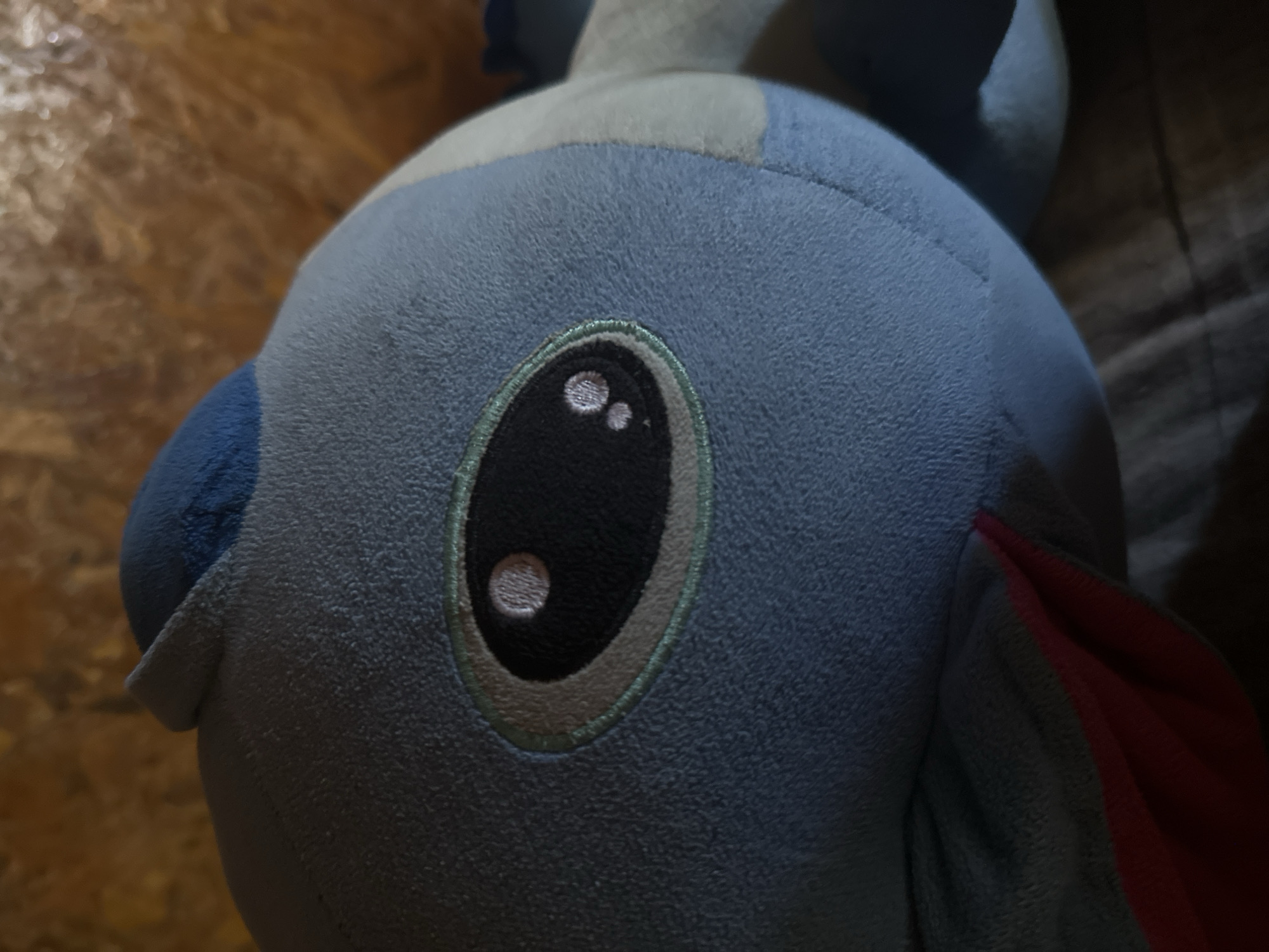The image shows a close-up of a light blue plush toy, possibly a creature with large, dark eyes outlined in light green. The toy has a rounded head with a darker blue section on top and what appears to be a pink or red mouth or fin area peeking out from the bottom. The background is blurred and consists of a textured surface in warm, golden-brown tones, which could be wood or a similar material. The toy is resting on a surface covered with a fabric that has a visible weave, appearing to be a neutral color like brown or gray. There is no visible indication of the city of Galiche, Bulgaria in the image.