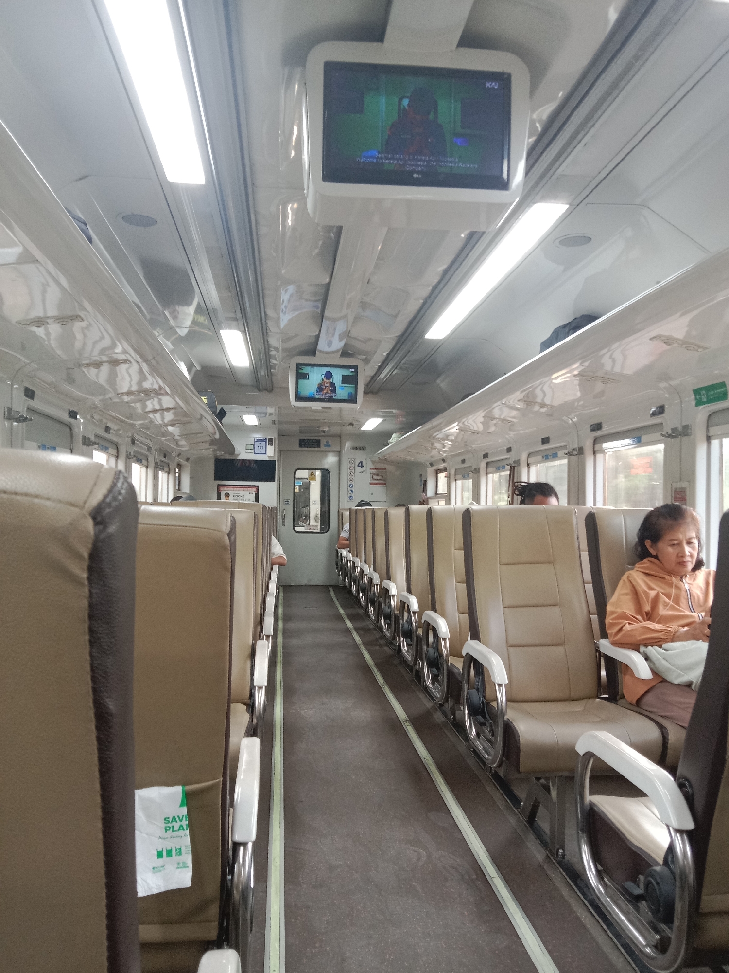The image captures the interior of a train carriage in Madiun, Indonesia. The carriage features rows of tan-colored seats with silver armrests. The aisle is carpeted in gray with a yellow stripe. Overhead, fluorescent lights illuminate the space, and two small televisions are mounted on the ceiling. One television displays a person in a red jacket, and the other shows a screen with text. A woman in an orange hooded jacket is seated on the right side of the train, facing forward. A white paper bag with green text that says "SAVE PLAN" is attached to the back of a seat on the left side. Several passengers are visible, some seated, and others partially obscured. The overall environment is that of a functional, modern train carriage.
