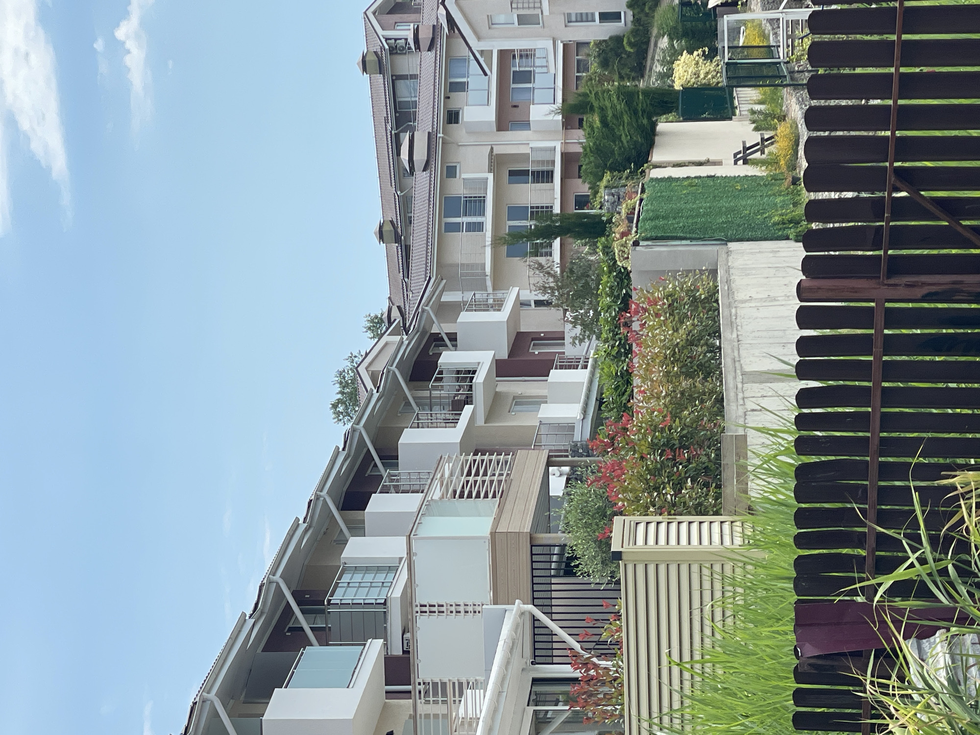 This is a medium shot, eye-level, vertical, outdoor, daytime photograph taken in Sochi, Russia. The image features a row of modern, multi-story residential buildings on a hillside. The buildings have light-colored facades, white trim, and dark brown tile roofs with visible chimneys. Balconies with glass railings and white vertical accents are present on multiple levels. In the foreground, there is a dark brown picket fence and tall green grass. Behind the fence, there is a beige louvered structure and vibrant green foliage with some red-tipped leaves. The sky is a pale blue with scattered white clouds. The buildings appear to be part of a resort or residential complex, likely situated near the coast given the sunny weather and lush vegetation, common characteristics of Sochi. No people are visible in the frame.