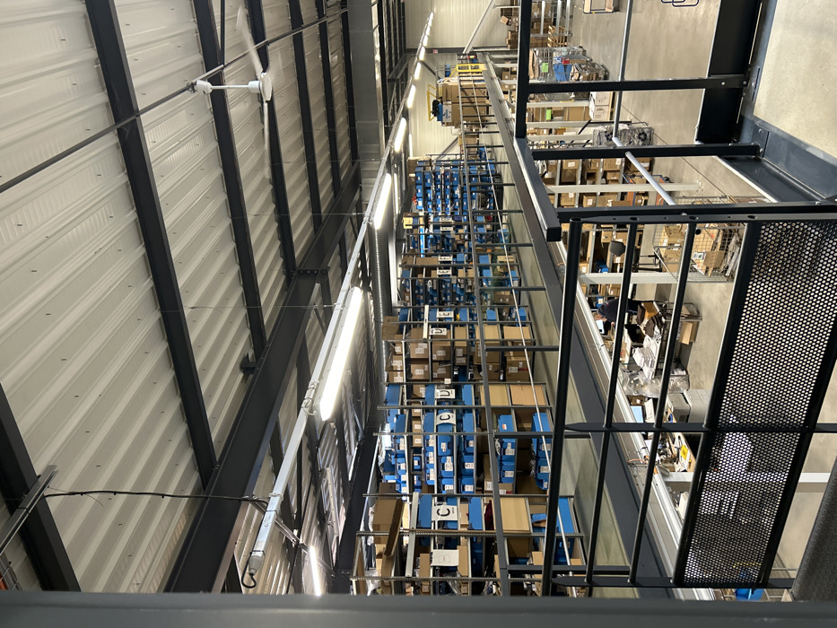 The image displays the interior of a multi-level industrial warehouse or distribution center located in Reichstett, France. The facility features a high, corrugated metal ceiling, light in color, supported by dark gray I-beams. Multiple illuminated fluorescent tube light fixtures are suspended from the ceiling, alongside at least one large, multi-bladed ceiling fan rotating in the upper right quadrant.
An extensive mezzanine level spans the middle and upper left portions of the scene, equipped with rows of gray metal shelving. These shelves are filled with numerous items, primarily contained within uniform blue plastic bins and various cardboard boxes. Several blue bins display white labels with single capital letters, such as "C" and "D." A dark gray metal railing, composed of horizontal bars and a lower perforated metal panel, encloses the edge of the mezzanine, overlooking the lower level.
On the lower level, directly beneath the mezzanine, a workstation area is visible. One individual, wearing a dark garment, is seated at a desk, oriented away from the camera, seemingly engaged with a computer monitor and associated paperwork. The desk area contains office equipment including printers and stacked documents. Further into the lower level, additional storage racks, multiple mobile carts (some metal, some blue), and stacked boxes are present. The floor on this lower level is a polished concrete surface. A light-colored wall or large door is visible in the background on the lower right.