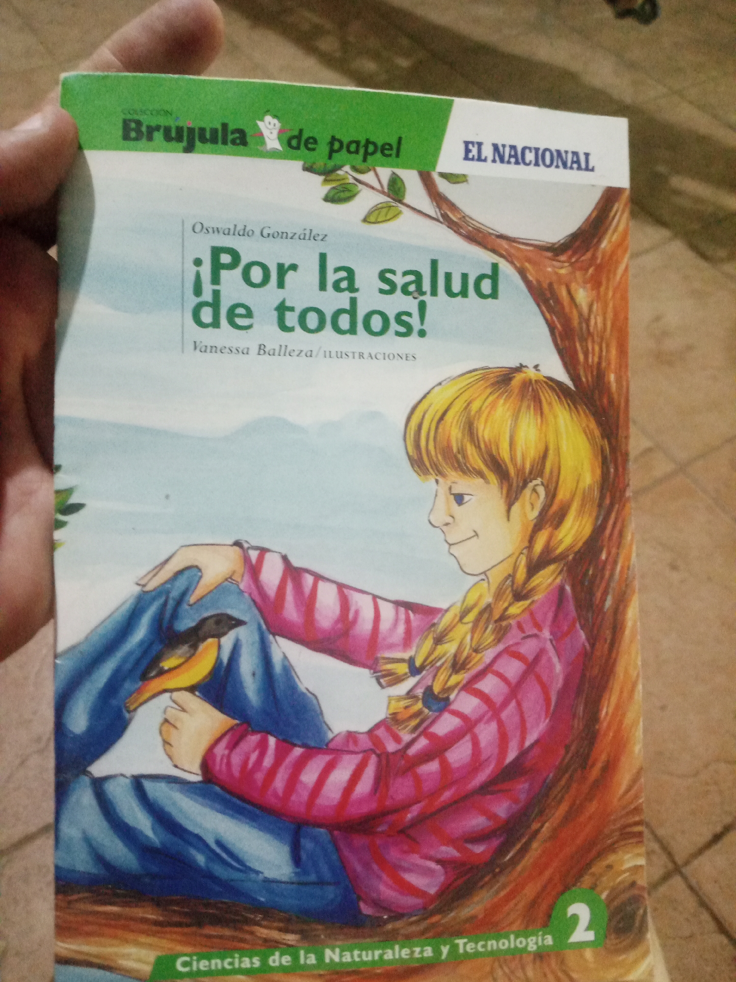 The image is a close-up of a book cover held by a person's hand. The book is titled "¡Por la salud de todos!" (For the health of all!) and features an illustration of a young blonde girl with braided hair sitting by a tree, with a small bird perched on her lap. The book cover also displays the publisher's name "EL NACIONAL" and the collection "Brujula de papel". The bottom of the cover reads "Ciencias de la Naturaleza y Tecnología 2", indicating it's part of a science and technology series. The background suggests an indoor setting with a tiled floor. There is no specific visual information within the image that would allow for its location to be identified as Santa Ana, Venezuela.