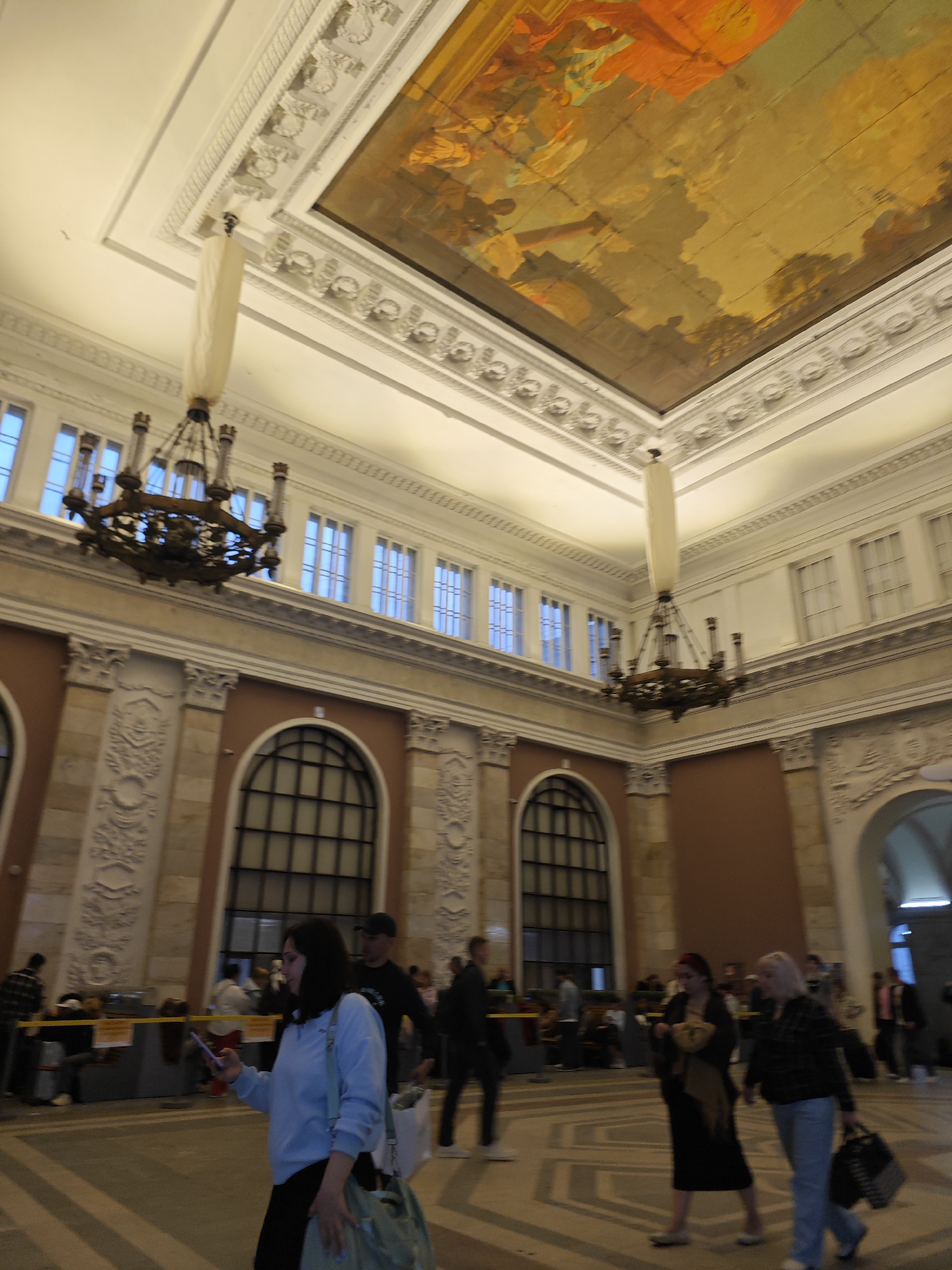 This is an indoor shot of a grand, ornate building interior, likely a train station or public hall, in Saint Petersburg, Russia. The architecture features high ceilings with elaborate decorative molding and a large, vibrant painted mural depicting a classical or allegorical scene across the upper portion of the ceiling. Two large, dark, multi-tiered chandeliers hang from the ceiling, their ornate metalwork casting a dramatic shadow. The walls are adorned with pilasters, decorative plasterwork, and large arched windows with dark frames.
In the foreground, a woman in a light blue sweatshirt and dark pants is walking to the left, looking down at her phone and carrying a light green bag over her shoulder. Behind her, a man in a dark t-shirt and black pants is walking in the same direction, carrying a white tote bag. Further in the background, several other people are visible, some walking and others gathered near a yellow barrier, suggesting a queue or waiting area. The floor is tiled with a geometric pattern. The overall impression is one of a well-maintained, historic public space.