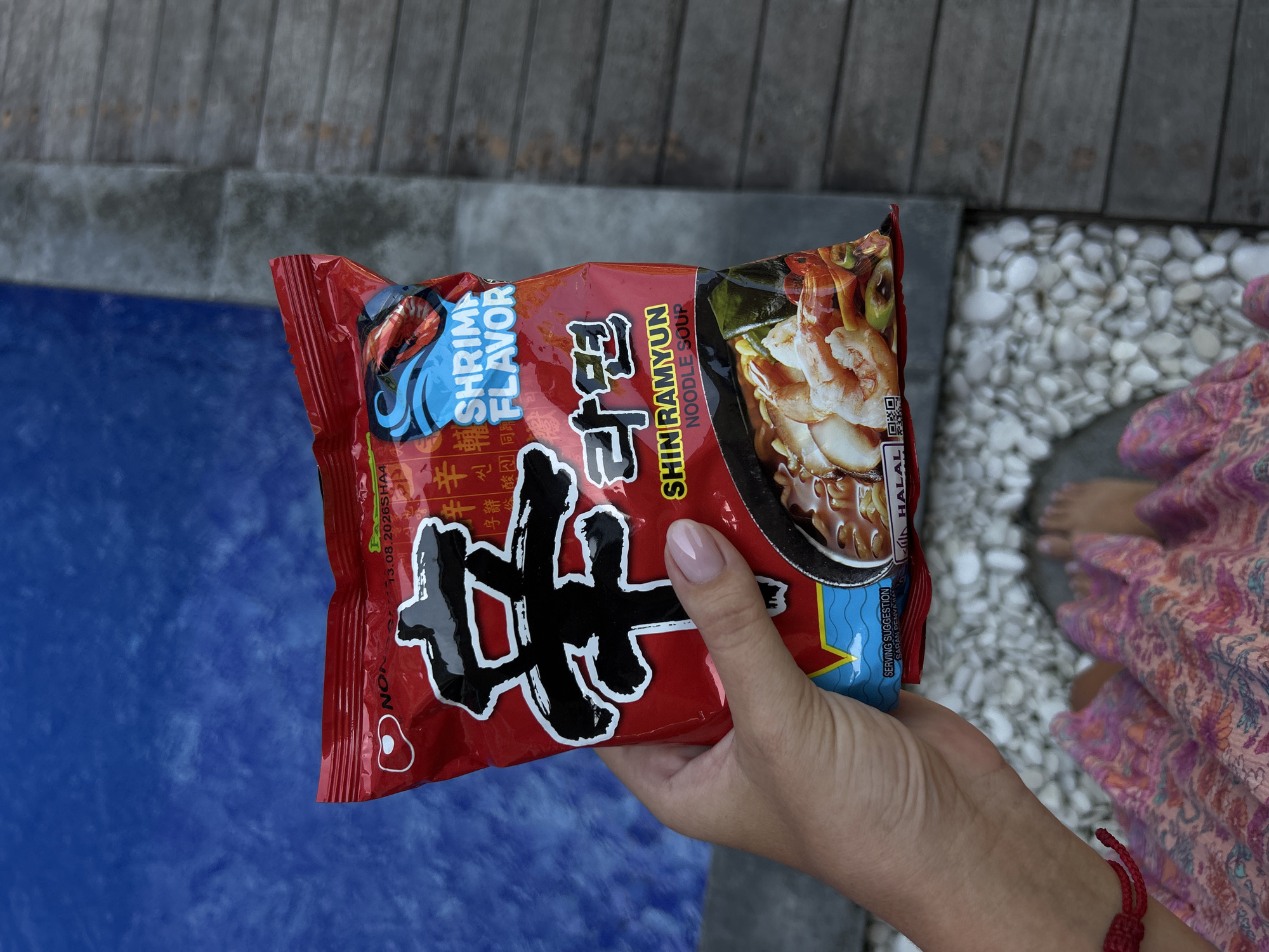 A person's left hand, with light skin and a pale pink manicure, holds a red and black package of "Shin Ramyun Noodle Soup Shrimp Flavor" ramen. The package is positioned in the foreground, slightly angled. The background shows a blue tiled swimming pool on the left and a wooden deck on the right, characteristic of outdoor spaces in tourist areas like North Kuta, Indonesia. White pebbles are scattered on the ground between the deck and the pool. A portion of the person's legs, clad in a pink patterned dress, is visible at the bottom of the frame, along with their bare feet. The overall scene suggests a casual, possibly vacation setting.