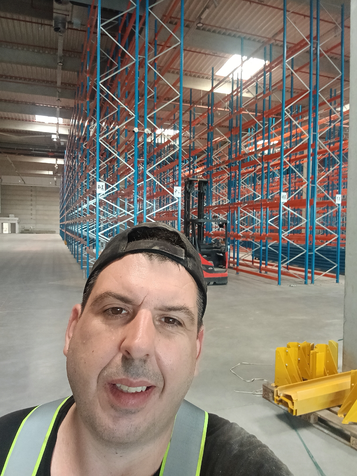 A man in a black baseball cap and a reflective vest is taking a selfie in a large, empty warehouse. The warehouse has tall, blue and orange metal shelving units lining the walls, stretching back into the distance. A red forklift is parked in the background, to the right of the man. Yellow metal pieces are stacked on pallets on the right side of the image. The floor is concrete, and the ceiling has exposed metal beams and some skylights. The man's face is close to the camera, and he appears to be smiling. The overall impression is of an industrial setting, possibly a distribution center or storage facility.