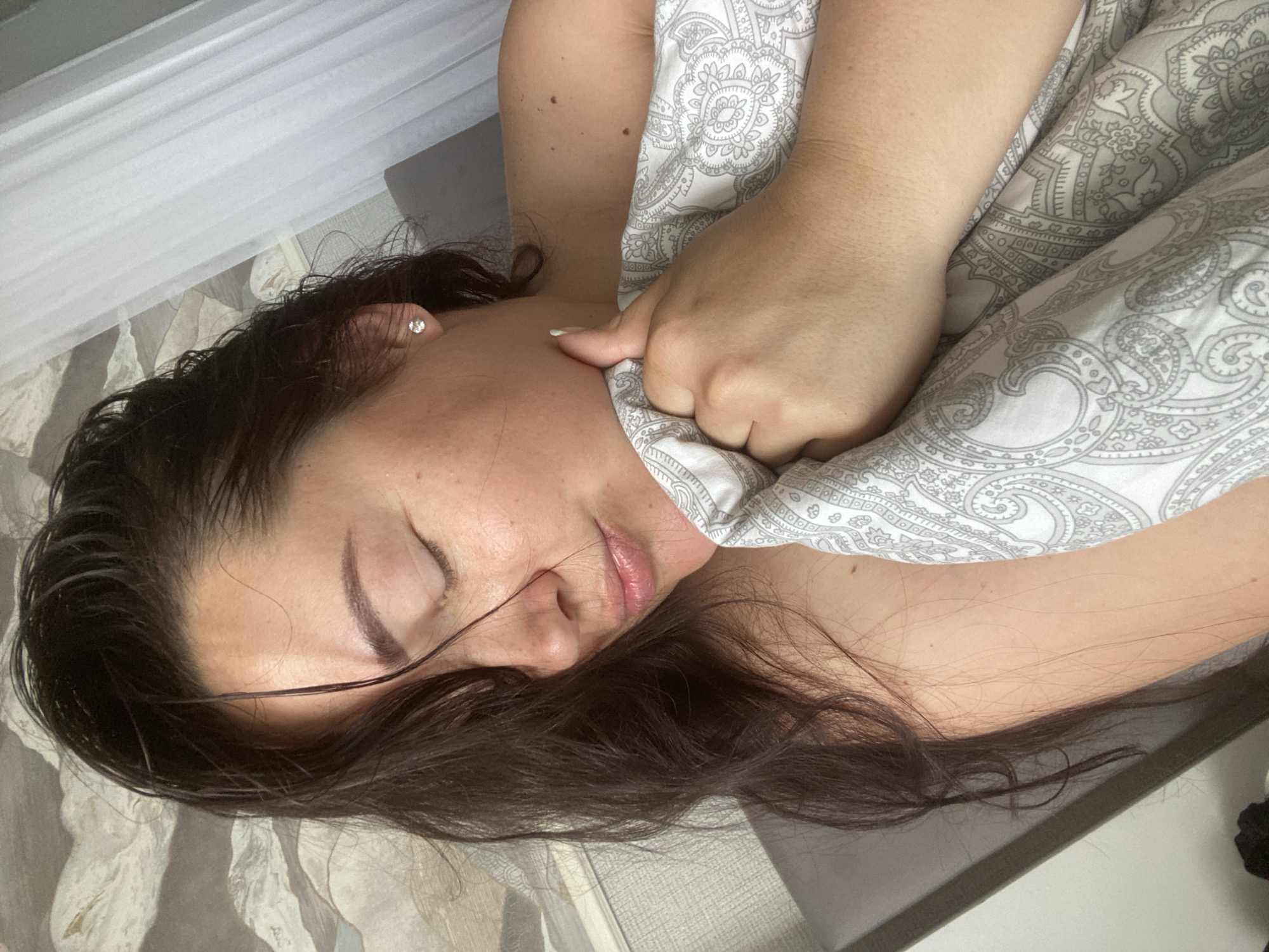 A woman is lying in bed, holding a patterned blanket up to her chin. Her eyes are closed, and her head is tilted slightly to the side. She has dark, wet hair and is wearing a single stud earring. The background shows a grey headboard and a white curtain. The image appears to be taken indoors, likely in a bedroom. There is no visible indication of the location being Sochi, Russia.