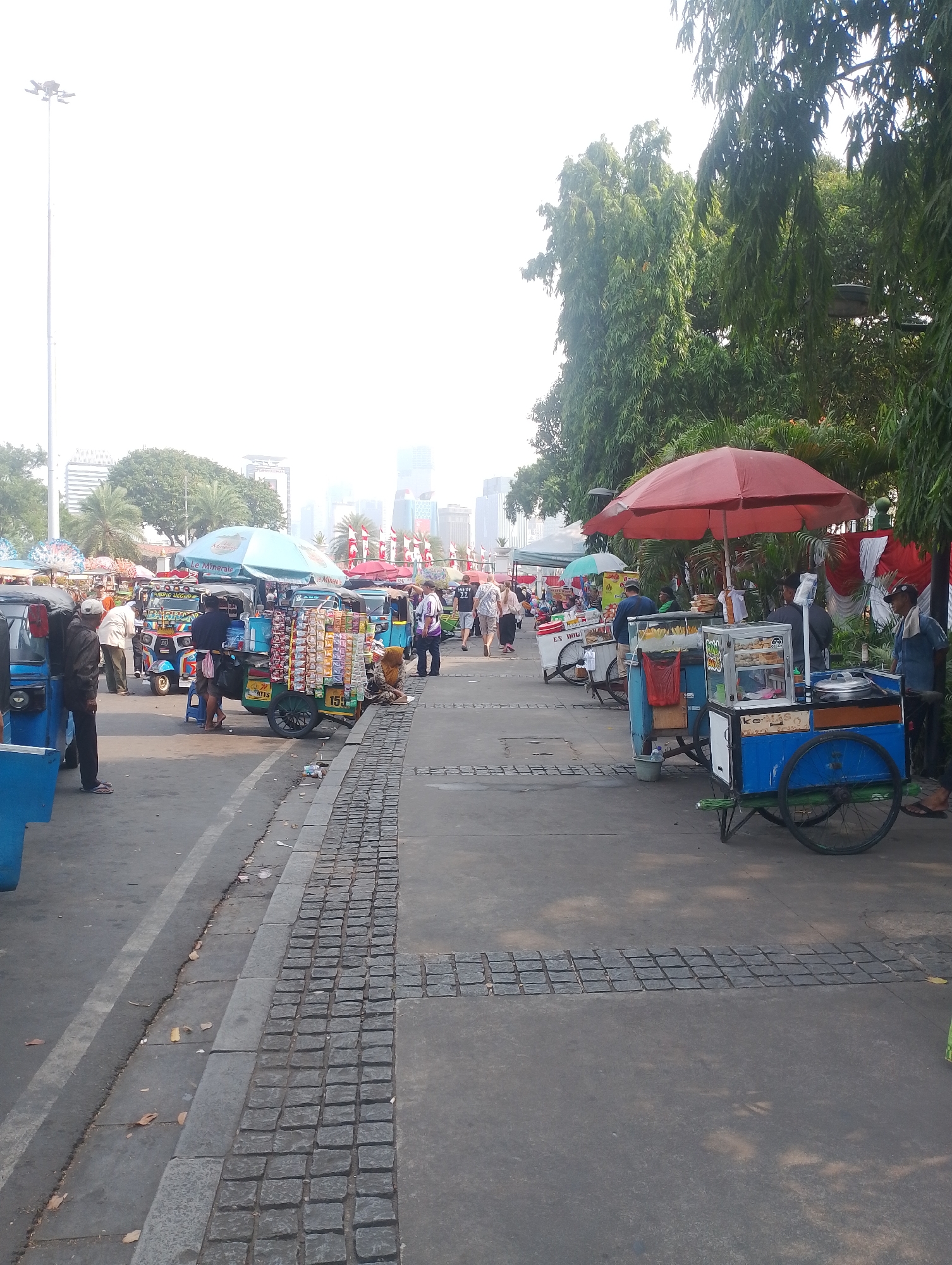 The image captures a bustling street scene in Central Jakarta, Indonesia, likely near Monas National Monument, a prominent landmark in the city. The foreground is dominated by a paved walkway lined with food carts and people. A blue food cart with a red umbrella shades a vendor preparing food, while another cart displays a variety of snacks. To the left, several blue and yellow auto-rickshaws are parked, some with passengers. In the background, lush green trees and modern skyscrapers suggest the juxtaposition of nature and urban development. Several individuals are seen walking and interacting, some browsing the food stalls and others conversing. The overall atmosphere is that of a lively market, with the sunlight casting shadows on the ground, indicating it's likely daytime.
