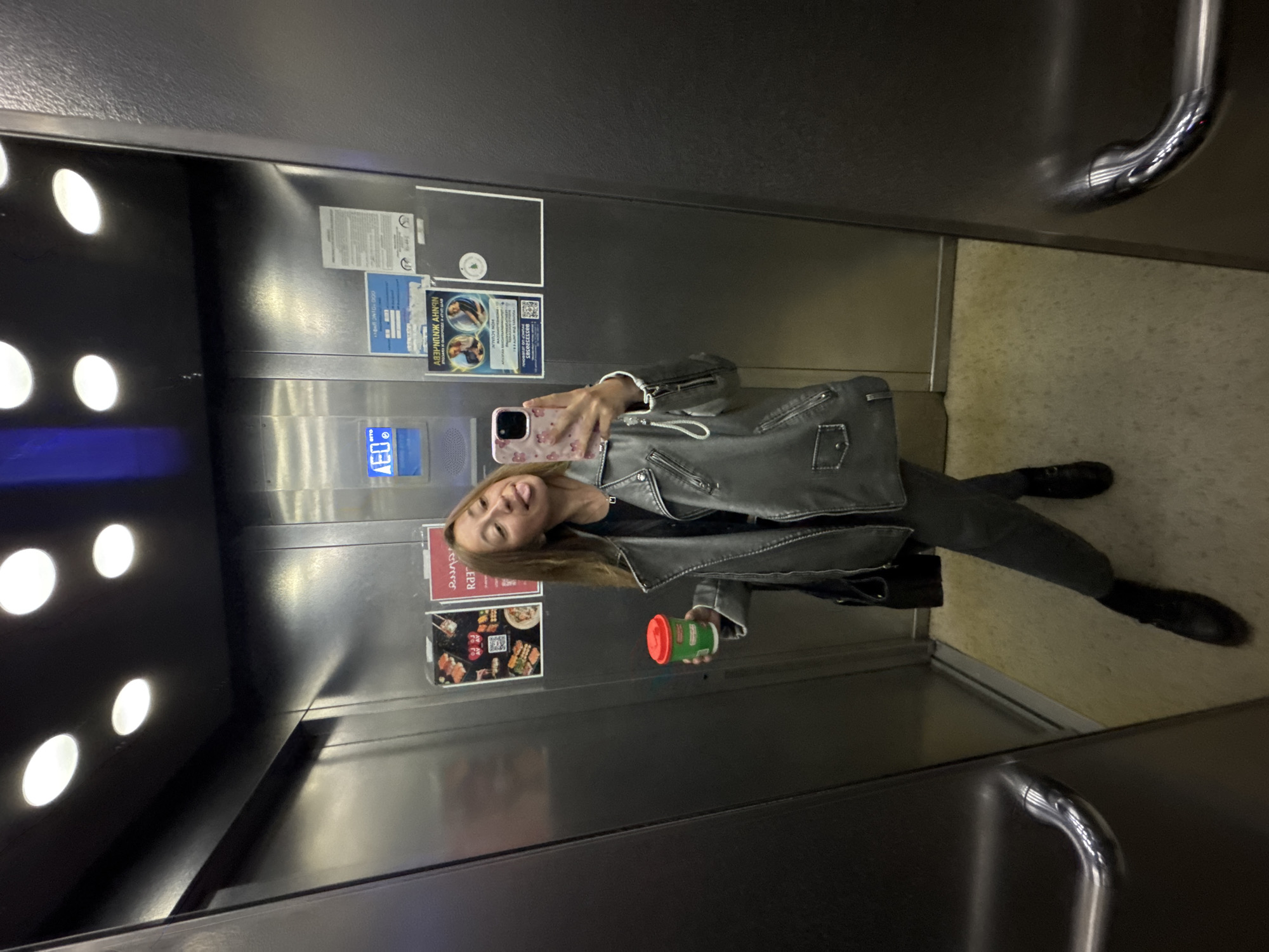 The image shows a woman in an elevator. She is holding a phone in one hand and a green and red container in the other. The elevator has advertisements and informational posters on the walls. The woman is wearing a gray leather jacket and dark pants. The location is likely Chaikovsky, Russia based on the text on the posters in the elevator.