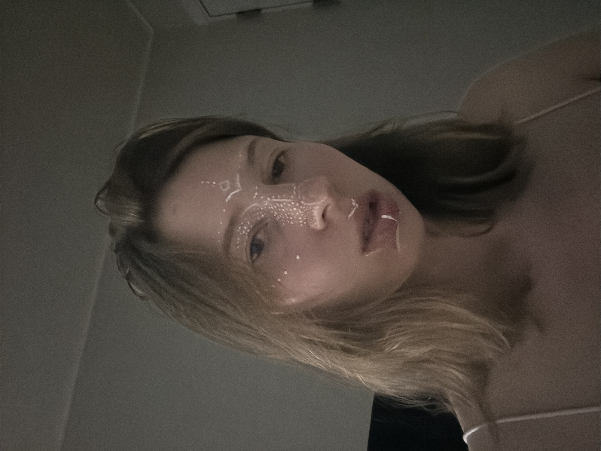 A young person with blonde hair, facing forward and slightly to the left, is the subject of this close-up portrait. Their face is adorned with white, intricate face paint that extends from their forehead down their nose and across their cheeks. The person is wearing a thin-strapped top. The lighting is dim, casting shadows and creating a moody atmosphere. The background is a plain, dark wall, suggesting an indoor setting. No specific landmarks of Dongducheon-si, South Korea are visible in this image.