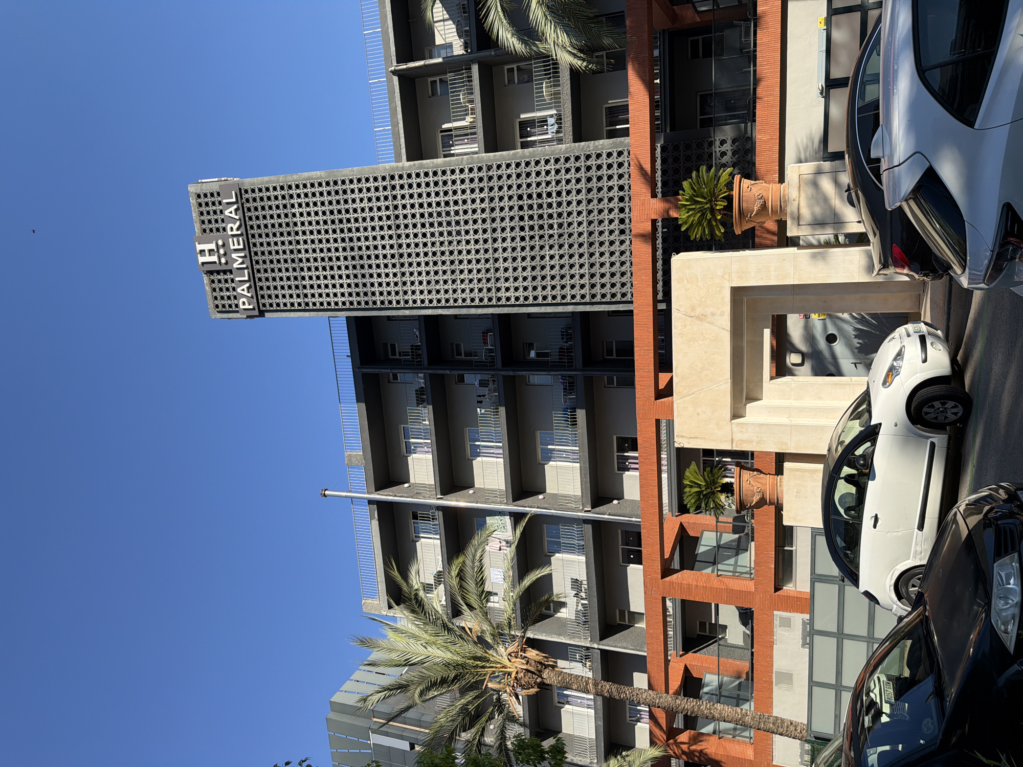 The photograph captures a street-level view of the Hotel Palmeral in Benidorm, Spain. The hotel features a distinctive tall tower with a patterned facade and the "H PALMERAL" sign at its apex. Below the tower, the hotel building itself is visible, characterized by multiple floors with balconies and windows. The facade of the lower section of the building is orange brick with arched openings. In the foreground, several cars are parked along the street, including a white compact car and a black sedan. Lush green palm trees are also present, adding to the tropical atmosphere. The sky is clear and blue, indicating a sunny day. The scene appears to be a typical urban streetscape in a Spanish coastal city.