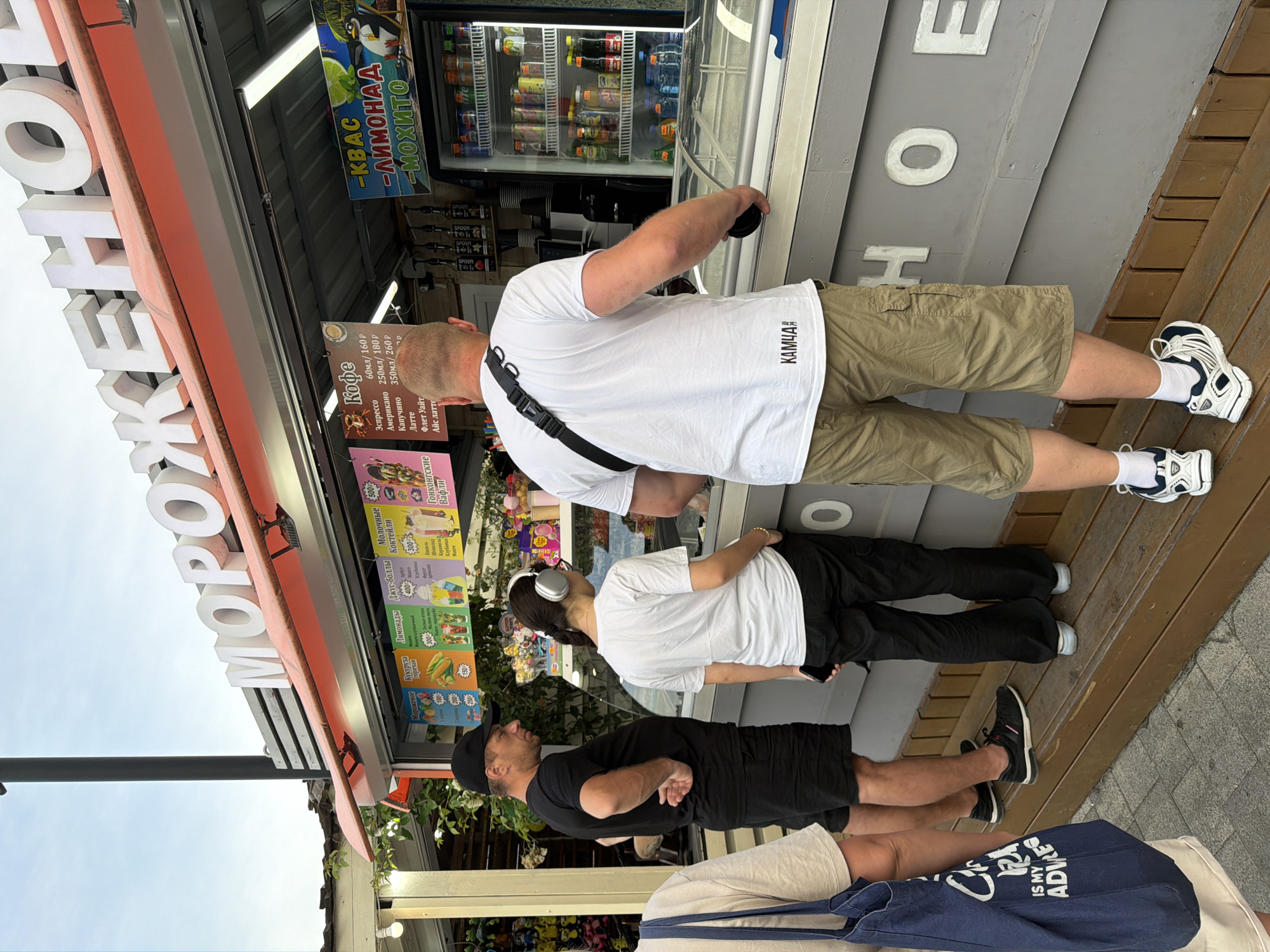 A group of people are standing and sitting in front of an outdoor kiosk selling ice cream and drinks. The kiosk has large, white, three-dimensional lettering on its facade that reads "МОРОЖЕНОЕ" (ice cream). There are colorful signs advertising various ice cream flavors, lemonades, and coffee drinks with prices listed in rubles. A refrigerator filled with bottled drinks is visible behind the counter.
One man, wearing a white t-shirt and khaki shorts, is holding a black cup and looking towards the kiosk. Another man, in a black t-shirt and black shorts, is wearing a black baseball cap and looking at the man in the white t-shirt. A third person, wearing a white t-shirt and black pants, has headphones on and is looking up at the kiosk. A fourth person, wearing light-colored shorts, is partially visible sitting in the foreground, with a blue tote bag at their feet.
The scene appears to be set outdoors during the daytime, with a bright sky visible. The kiosk is situated on a wooden platform, suggesting a public area like a boardwalk or street. The overall atmosphere seems casual, with people engaged in ordering or waiting for their purchases. The visible text includes names of drinks and prices, and a brand name "КАМЧА" on the white t-shirt. There's also text on the tote bag that reads "IS MY ADVICE." The location is identified as Sochi, Russia.