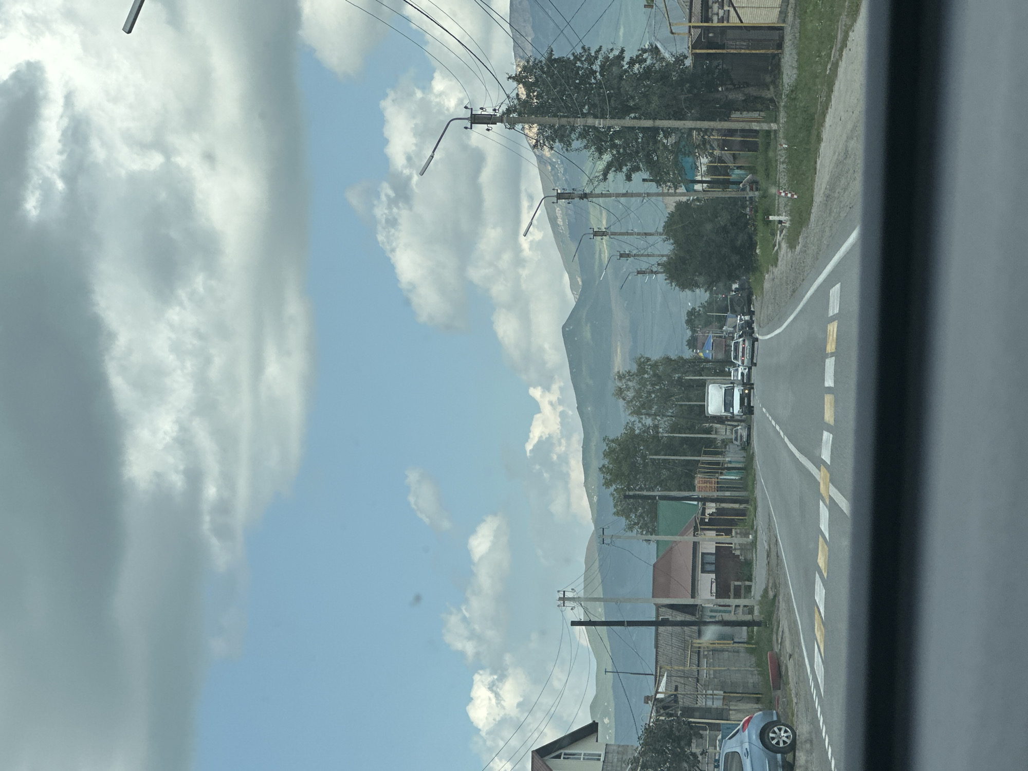 The image captures a street view in Zelenchukskaya, Russia, likely on a cloudy day. The perspective is from inside a vehicle, looking forward down a paved road. On the left side of the road, there is a blue car parked near a pedestrian crossing. The road itself is marked with white and yellow stripes, indicating a pedestrian crossing. Houses with various roof colors, including brown and green, line the right side of the street, interspersed with trees and utility poles carrying numerous wires. In the distance, a white truck and several other cars are visible on the road. Beyond the houses and trees, a range of green mountains forms the backdrop, under a sky filled with large, puffy white and gray clouds. The overall scene suggests a rural or semi-urban setting in a mountainous region.