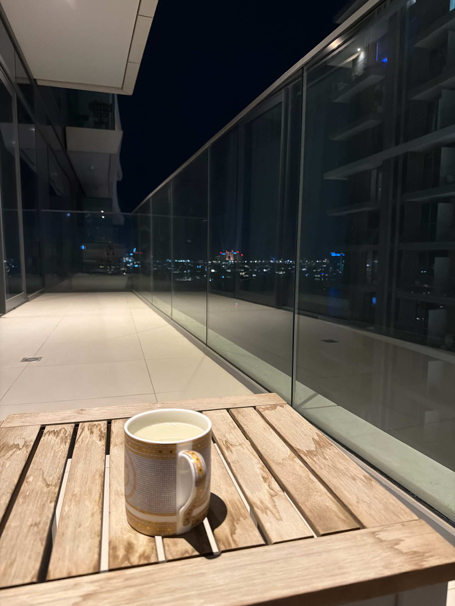 A mug of light-colored liquid sits on a wooden slatted table on a balcony at night. The balcony has a tiled floor and is enclosed by a glass railing. Beyond the railing, a city skyline is visible with numerous lights. The reflection on the glass indicates that the balcony is part of a modern building. The scene is likely in Dubai, United Arab Emirates, given the architectural style and urban density suggested by the skyline.
