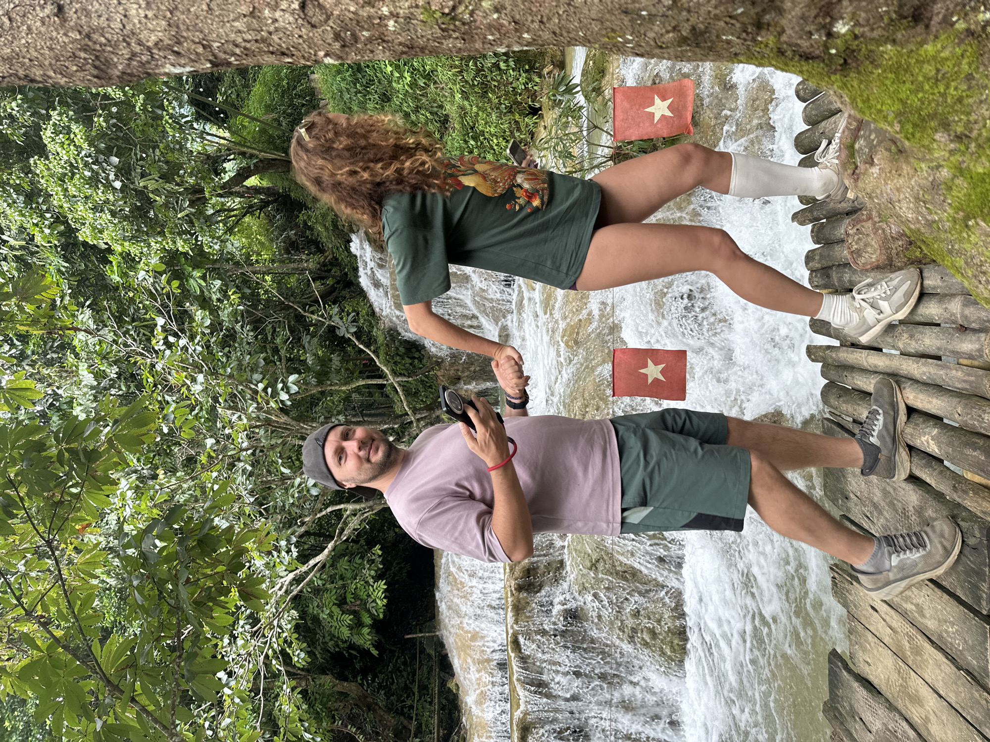 A man and a woman are walking hand-in-hand on a wooden walkway near a waterfall in Cổ Lũng Commune, Vietnam. The man is wearing a purple t-shirt, green shorts, and grey sneakers. He is holding a phone in his right hand. The woman is wearing a green t-shirt with a rooster design, white socks, and white sneakers. She has long, curly brown hair and is wearing a gold hair clip. Two red flags with white stars are visible behind them, suggesting a national or regional context. The environment is lush with green trees and vegetation, and the waterfall is flowing with murky water. The walkway appears to be constructed from logs and bamboo.