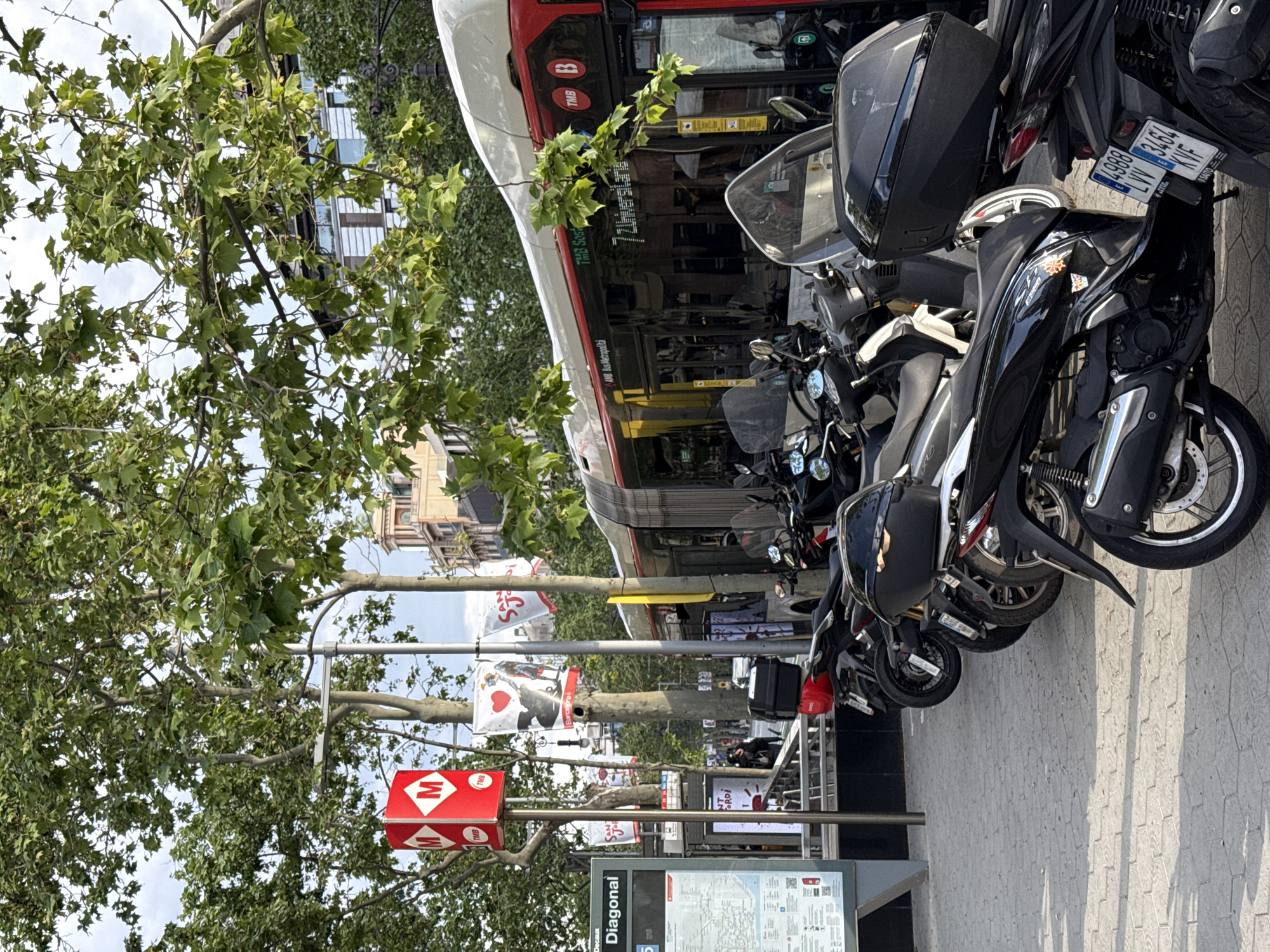 The image depicts a street scene in Barcelona, Spain. In the foreground, a "Diagonal" metro station sign is visible, along with a map display of the metro system. A red and white metro sign, marked with a stylized "M" logo, is also present.
A row of parked scooters and motorcycles lines the sidewalk. Adjacent to the scooters is a bus, specifically identified as a TMB Sost bus, number 72 Universitaria.
In the background, buildings can be seen, and a tree with green foliage partially obscures the view. The sky appears bright with some clouds visible. The scene appears to be taken during the daytime.