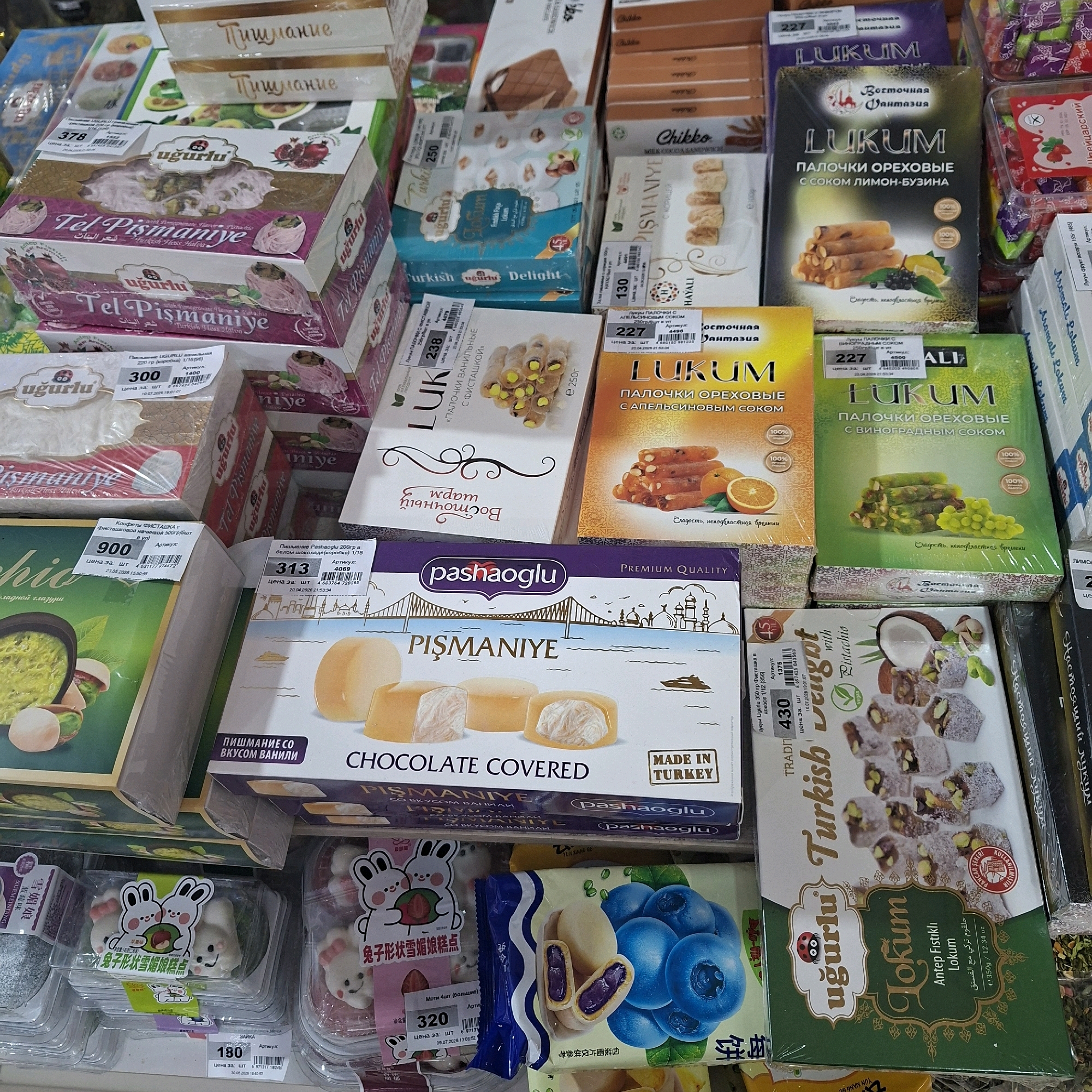 The image shows a retail display of Turkish confections, likely in a market or grocery store. The products are primarily boxes of "Lokum" (Turkish Delight) and "Tel Pismaniye," a type of shredded pastry. The display is densely packed, with multiple stacks of colorful boxes. Labels and prices are visible on many of the products, some in Russian and some in English, indicating a potential market in a Russian-speaking region. The lighting is even, suggesting an indoor setting with artificial illumination. There are no people visible in the image. The focus is on the product packaging, which features intricate designs and images of the confections, fruits like grapes and pomegranates, and various text elements.
