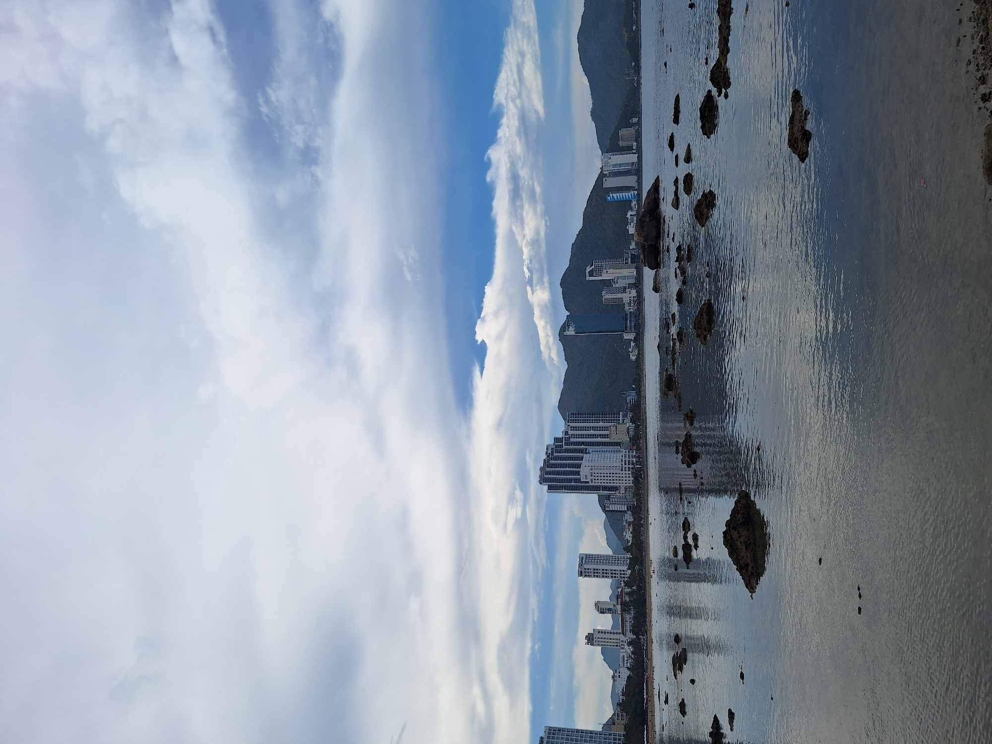 The image depicts a coastal cityscape in Nha Trang, Vietnam, under a cloudy sky with patches of blue. In the foreground, the shallow water of the sea is visible, with numerous dark, jagged rocks partially submerged. The water reflects the sky and the distant buildings, creating a slightly distorted mirroring effect. Along the coastline, a sandy beach is lined with numerous tall, modern buildings, primarily white, arranged in a dense formation. Behind the buildings, lush green mountains rise, forming a dramatic backdrop. The sky is filled with a mixture of white and grey clouds, with some areas revealing a bright blue sky. There are no people or animals clearly visible in the image. The overall scene suggests a serene, perhaps early morning or late afternoon, view of a developed coastal area.