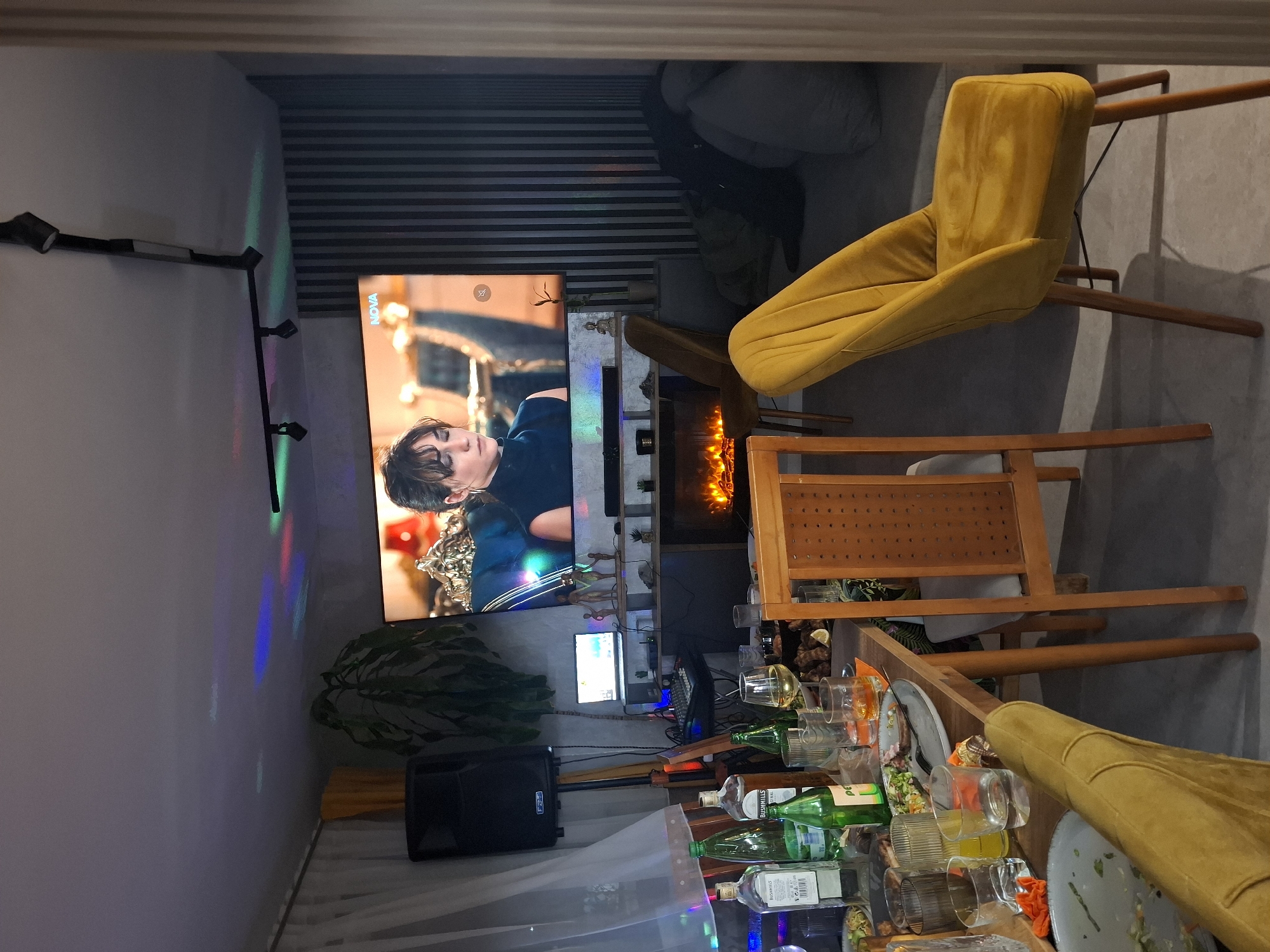 The image shows a cluttered living room interior. A large television displays a scene with a person in focus. A sound system sits on the floor near a plant. A shelf unit holds various decorative items, and an electric fireplace is integrated into the design. The dining table is laden with dirty dishes, empty bottles, and numerous glasses. Two yellow chairs are placed near the table. The location is unspecified, and no connection to Veliki Preslav, Bulgaria, is explicitly confirmed.