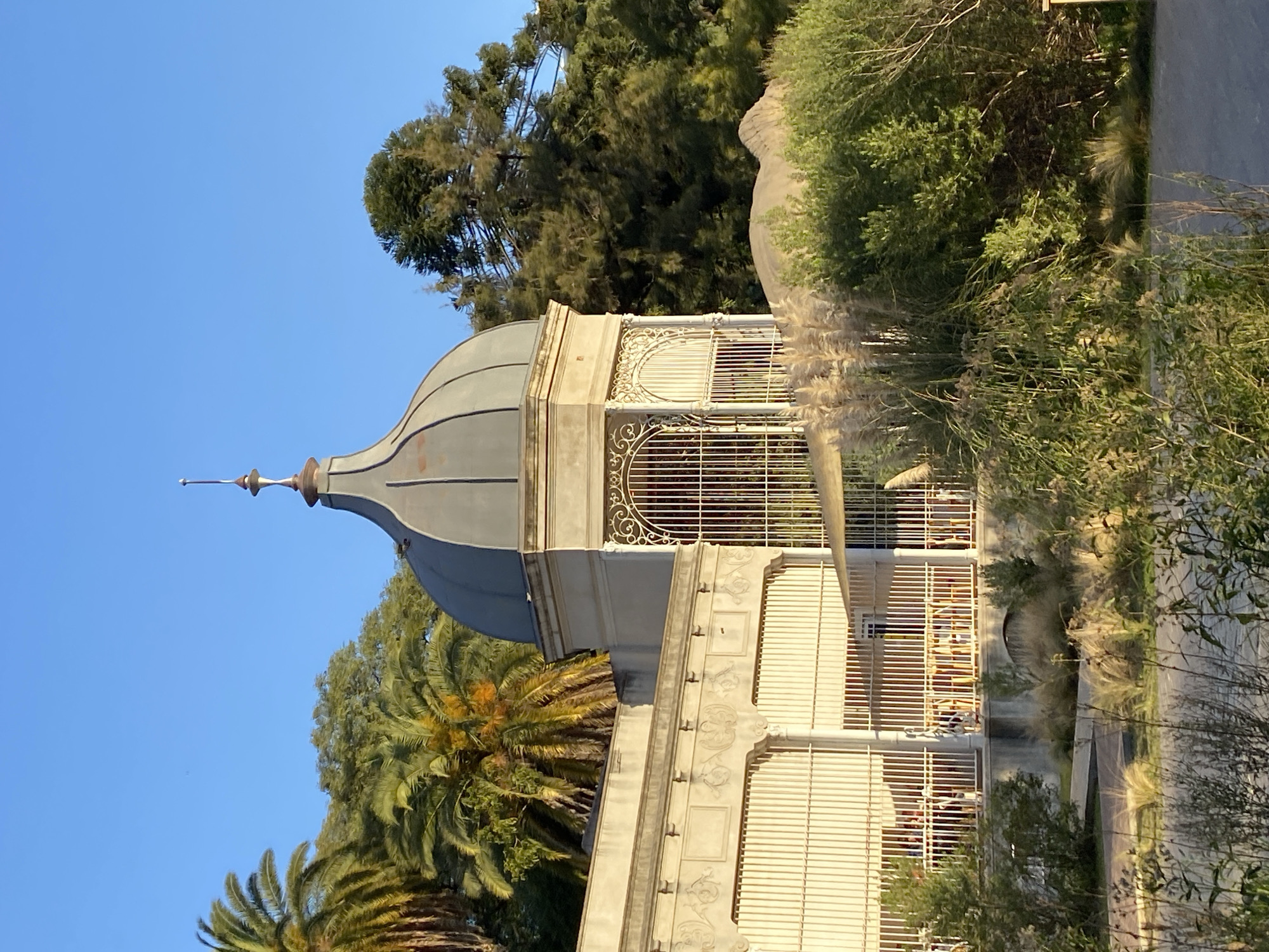 The image shows the dome and upper structure of the Pabellón de las Rosas, located in the city of Buenos Aires, Argentina. The pale building features a gray dome and ornate white metalwork. Trees and palm trees surround the building. The sky is blue. Some people are seated inside.