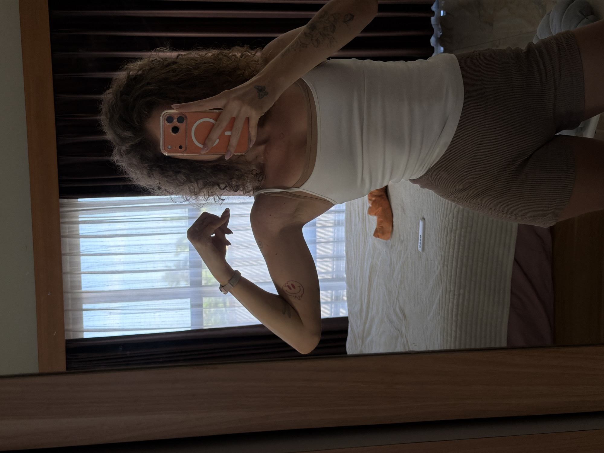 A young woman with curly brown hair takes a mirror selfie in what appears to be a bedroom. She is wearing a white tank top layered over a nude camisole and brown ribbed shorts. Her left arm is flexed, showing her bicep, and she has tattoos on both arms. She holds a smartphone in front of her face to take the photo. In the background, a bed with white and pink bedding is visible, along with sheer curtains and dark brown drapes. An orange object and a white object that looks like a remote control are on the bed. The room is lit by natural light coming through the window. The location is stated to be in the city of North Kuta, Indonesia.