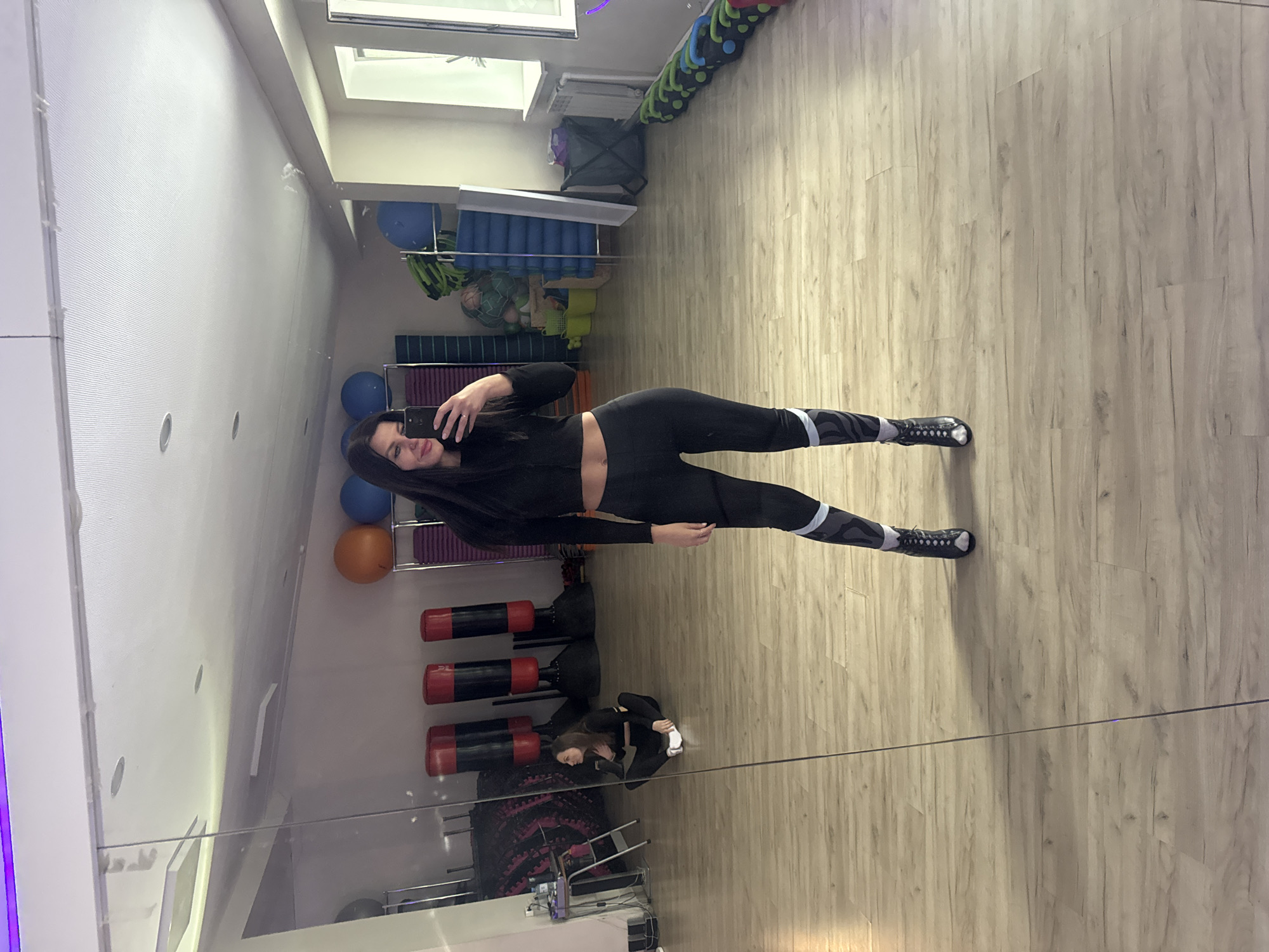The image shows a woman in a black crop top and leggings with a grey and white stripe taking a selfie in a gym. The woman is standing, facing the camera, with her phone in her hand. In the background, there are gym equipment such as exercise balls, foam rollers, resistance bands, and punching bags. The gym floor is light-colored wood. The reflection shows another person sitting down. The image was taken in a gym in Chaikovsky, Russia.