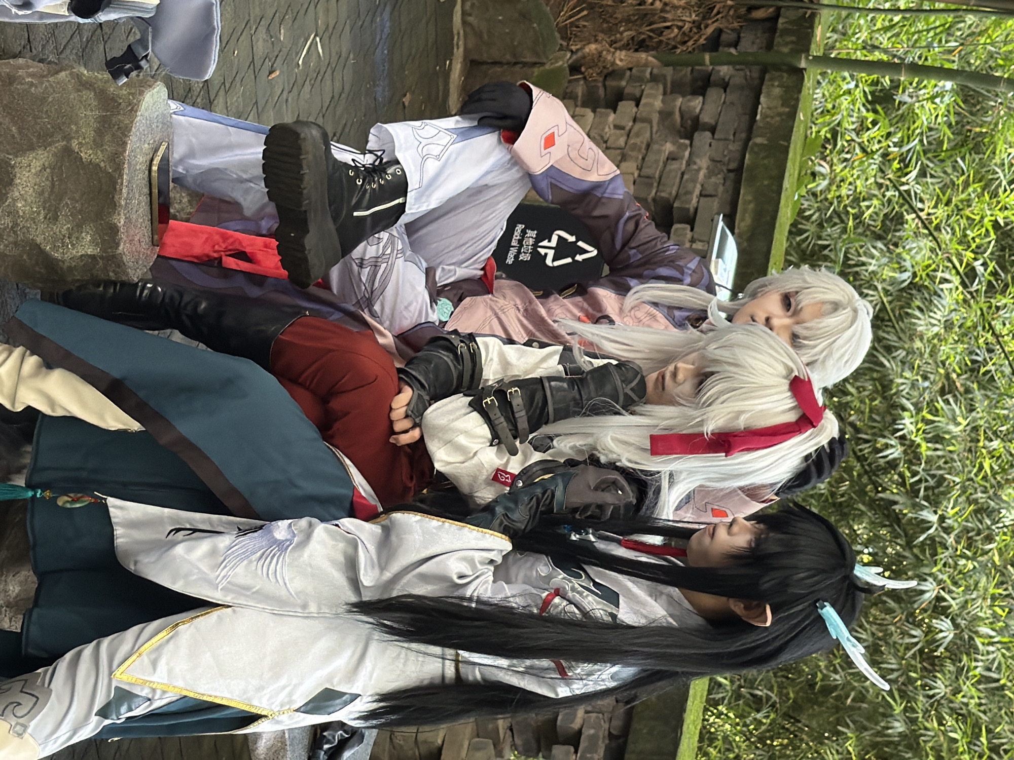 Three individuals dressed in cosplay attire are seated on a stone bench outdoors, with lush green bamboo and foliage forming the backdrop. A brick wall runs behind them.
The person on the left, wearing a dark purple and pink patterned garment with white pants and black boots, is leaning back with their head tilted slightly. They are wearing black gloves.
In the center, an individual with long, white hair tied with a red ribbon is wearing a white and red outfit with black leather accents, including gloves and boots. They are looking downwards.
On the right, a person with long black hair, adorned with light blue, wing-like accessories, is wearing a light gray garment with intricate designs and golden trim. They are wearing dark gloves.
In front of the seated individuals, a large, rough-hewn stone serves as a small pedestal, on which a smartphone is placed. To the left of the stone, a light blue bag is partially visible. A red cloth is draped over the stone bench.
The ground appears to be paved with interlocking stones or bricks. The overall scene suggests a cosplay event or gathering in an outdoor, natural setting. The location is identified as Yuzhong District, China.