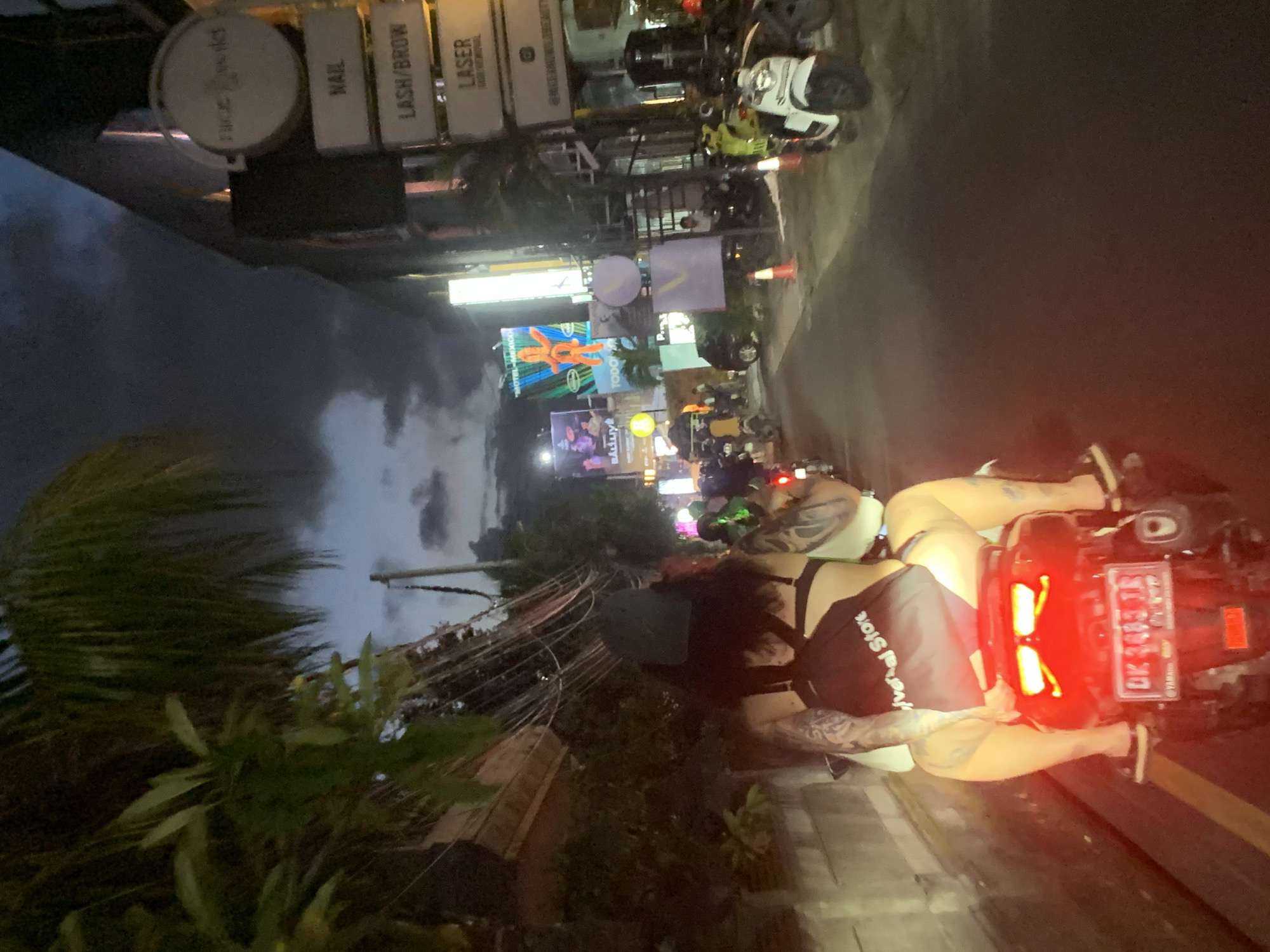 The image shows a street scene in North Kuta, Indonesia, at night. The street is lined with businesses offering services such as nail care, eyelash and eyebrow treatments, and laser treatments. Several motorbikes are parked along the street. A person on a motorbike is visible in the foreground, with their back to the camera. The license plate of the motorbike reads "DK 3483 TR." Various billboards and neon signs are visible in the background, including the name "BALLLIVE" and "TODO." Palm trees and other vegetation are present. The sky is visible in the background, suggesting it is either dusk or dawn. There is a store named "Verbal Store."
